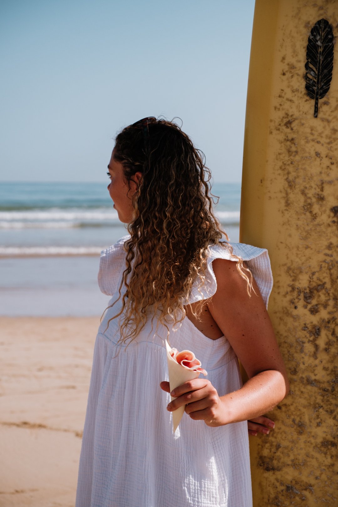 Femme tenue en robe blanche appuyée contre une planche de surf avec son cornet
