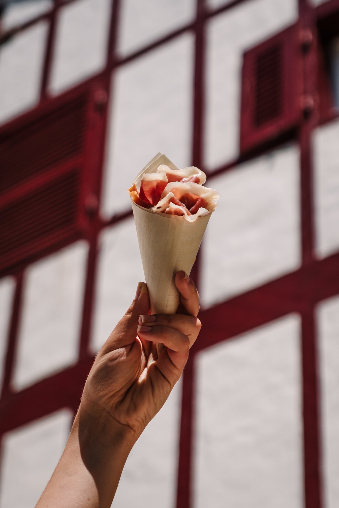 Main tenant un cornet de jambon cru devant une façade blanche aux volets rouges du Pays Basque