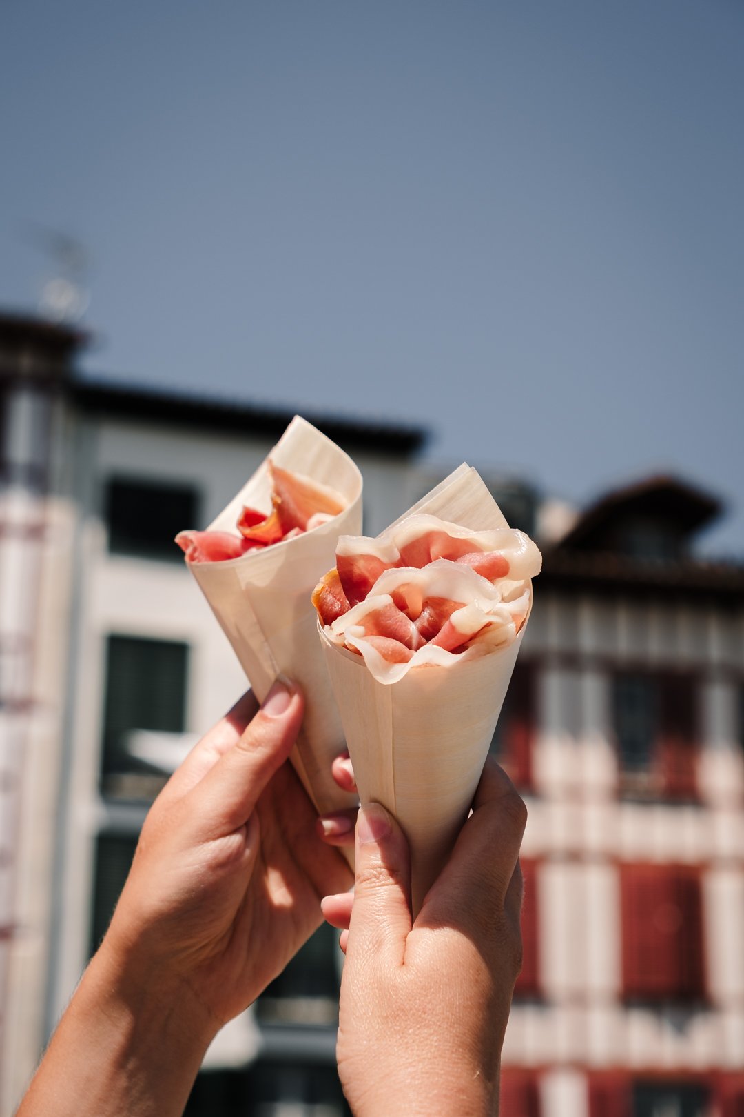 Deux cornets de jambon cru levés devant une maison traditionnelle à colombages à Bayonne