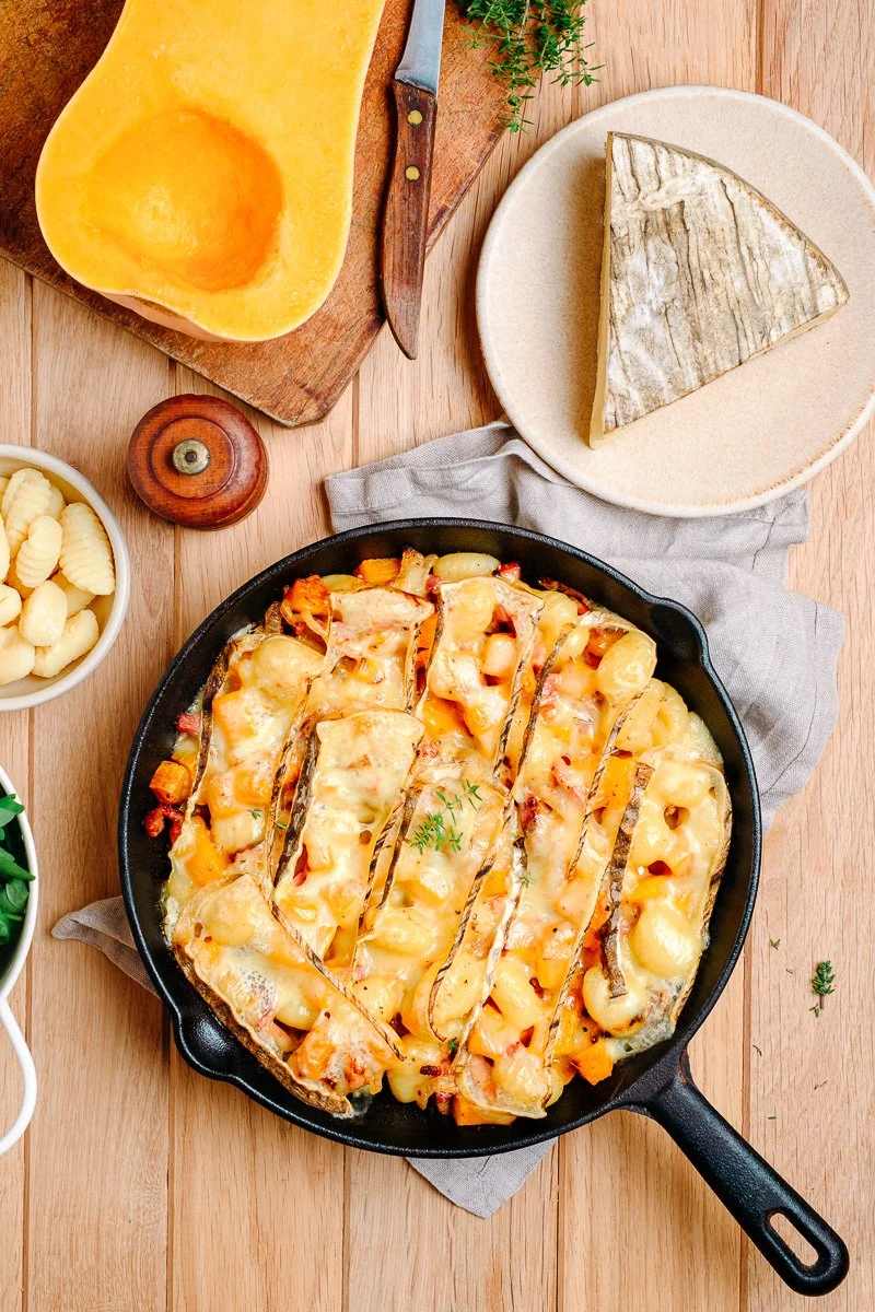 Gratin de pâtes avec des tranches de poisson gratinées au fromage, servi dans une poêle noire. Autour, un morceau de fromage, une cruche en bois, un bol de gnocchis, et une assiette avec du fromage.