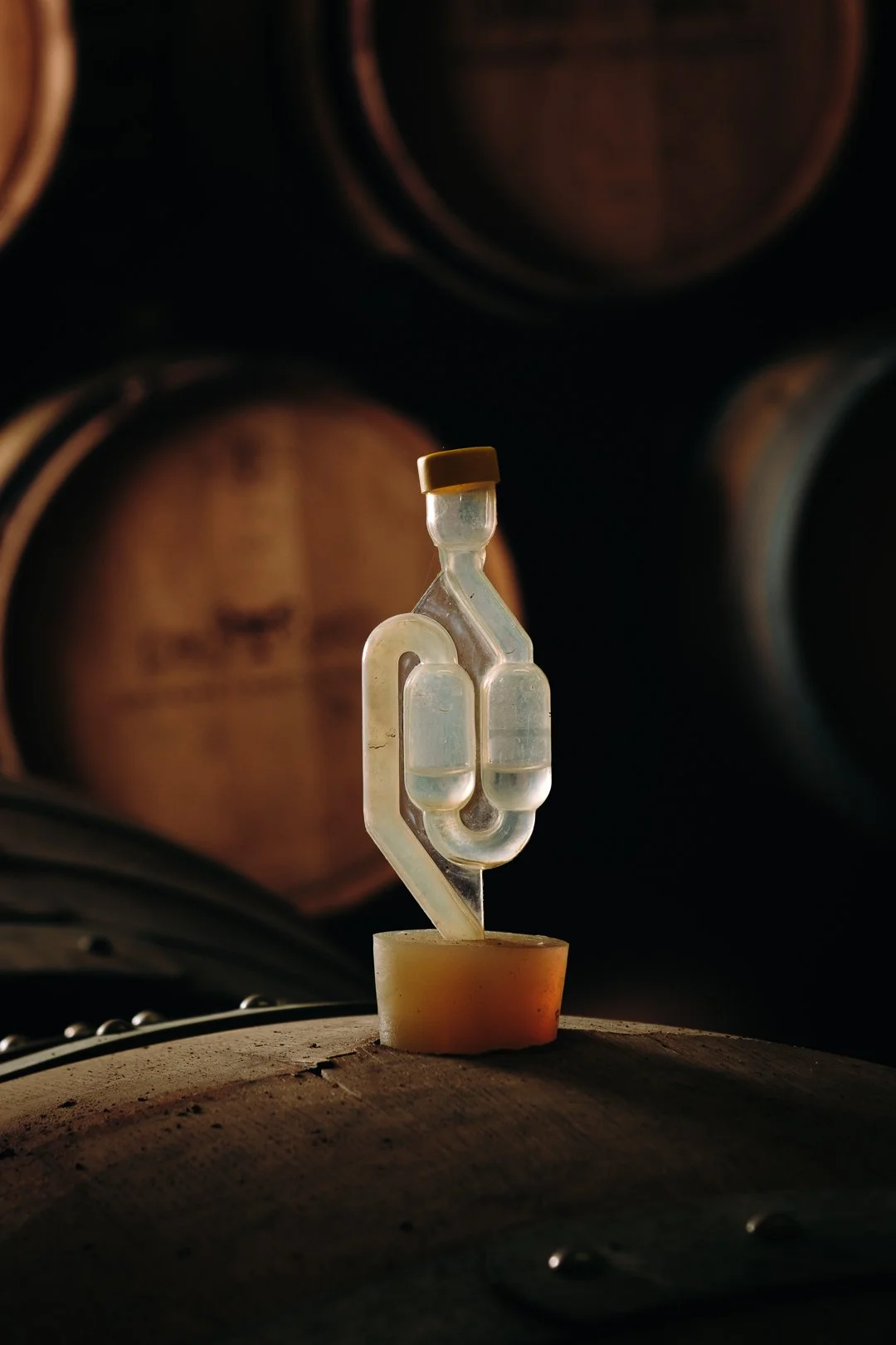 Une sculpture moderne en verre claire, représentant un flacon en forme de bouteille, placée sur un tonneau en bois dans une cave à vin ou un vignoble, avec des tonneaux en arrière-plan.