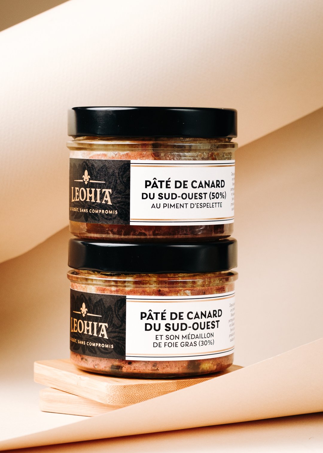 Deux pots de pâté de canard du Sud-Ouest empilés, le haut avec du piment d'Espelette, l'autre avec du foie gras, sur une petite planche en bois.