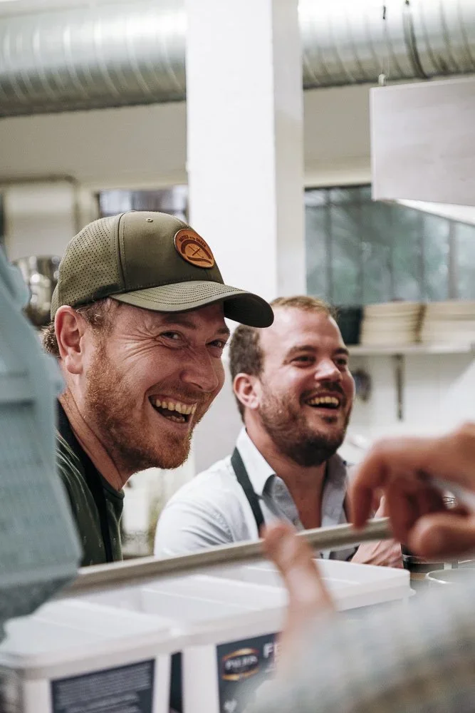 Deux artisans souriants en pleine préparation, reportage photographique immersif pour montrer l'humain derrière le métier.