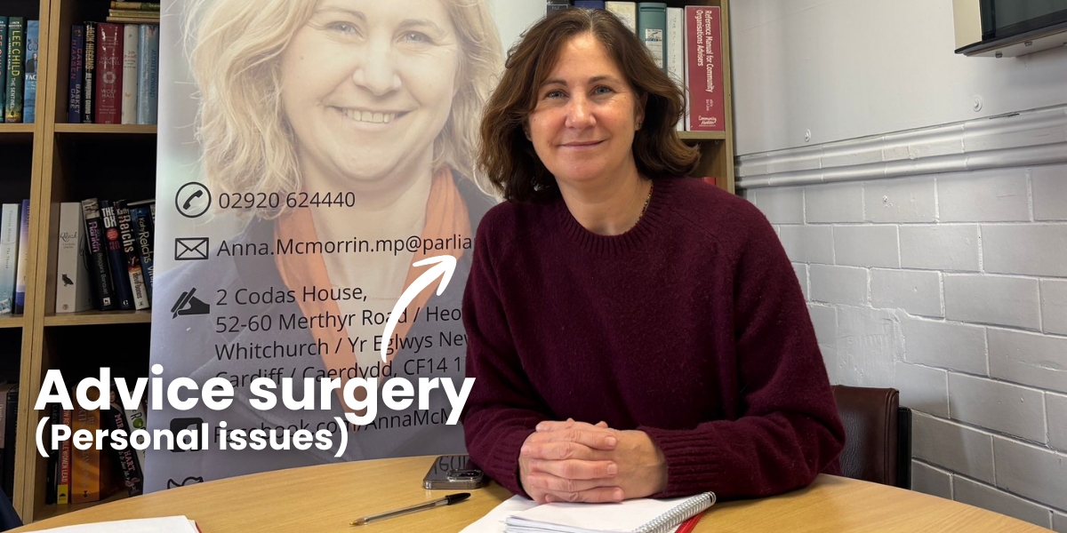 Anna McMorrin MP sitting at a desk with a notebook and pen, smiling in a room with book shelves and a poster behind her that shows her photo, contact information, and the words 'Advice surgery (Personal issues)'.