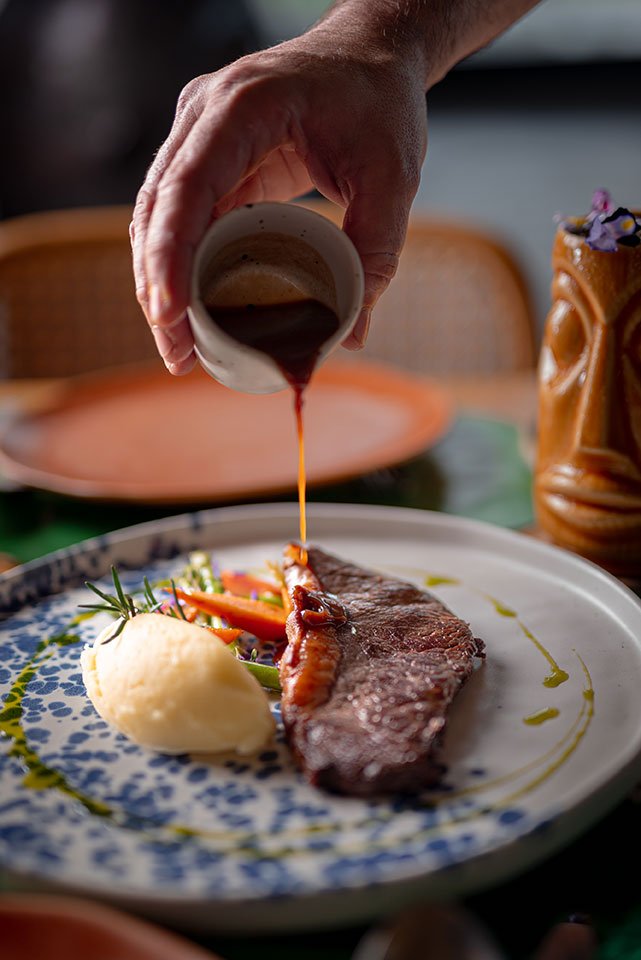 A hand pouring rich gravy over a perfectly cooked steak at Koa Dining, Tolani Koh Samui.