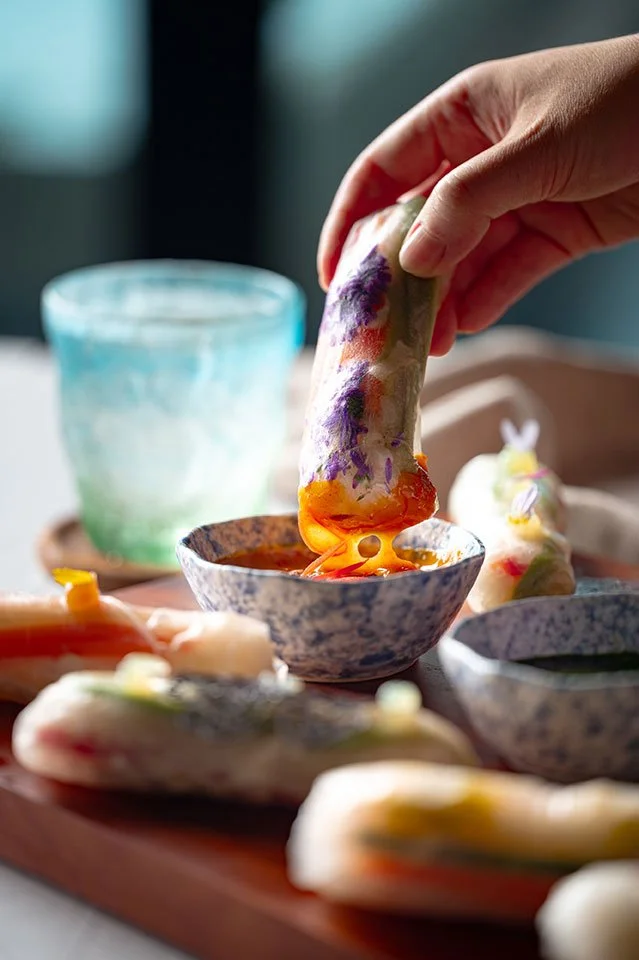A hand dipping a fresh spring roll into a flavorful bowl of sauce at Koa Dining, Tolani Koh Samui.