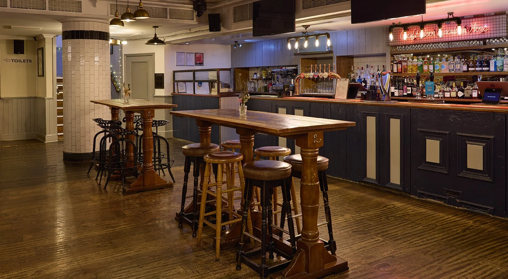 Interior of a cozy pub with wooden high tables and bar stools, a fully stocked bar in the background, and a white tiled pillar. Dimly lit with a warm atmosphere.