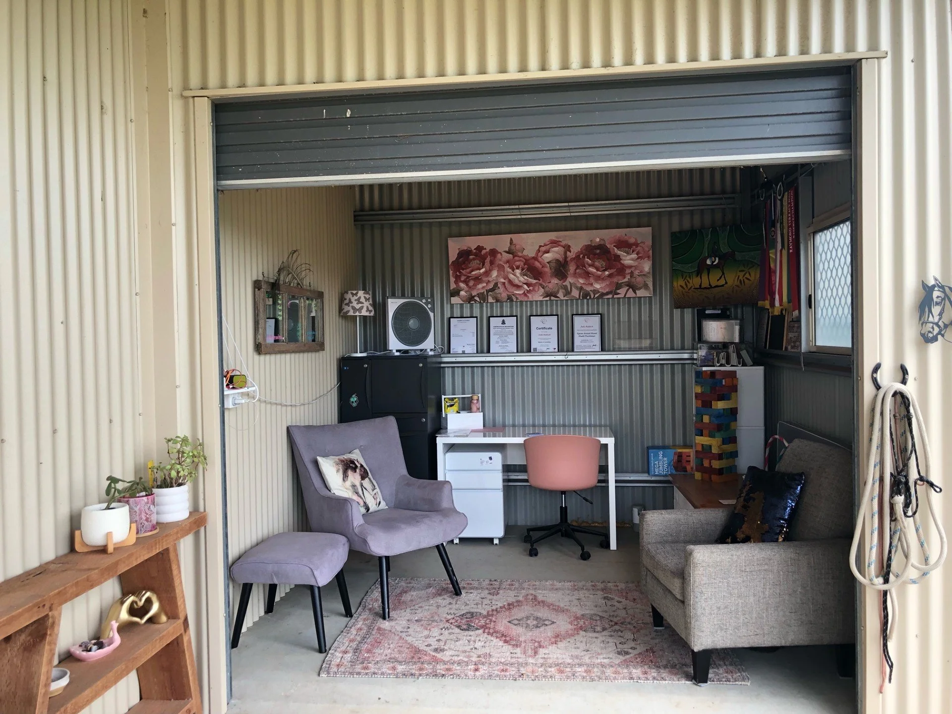 Interior of cozy room with beige corrugated walls, featuring armchair with pillow, grey sofa, small wooden table, a white desk with a pink rolling chair, shelves, plants, books, certificates, and decorative items. Stable Surroundings Equine Therapy.