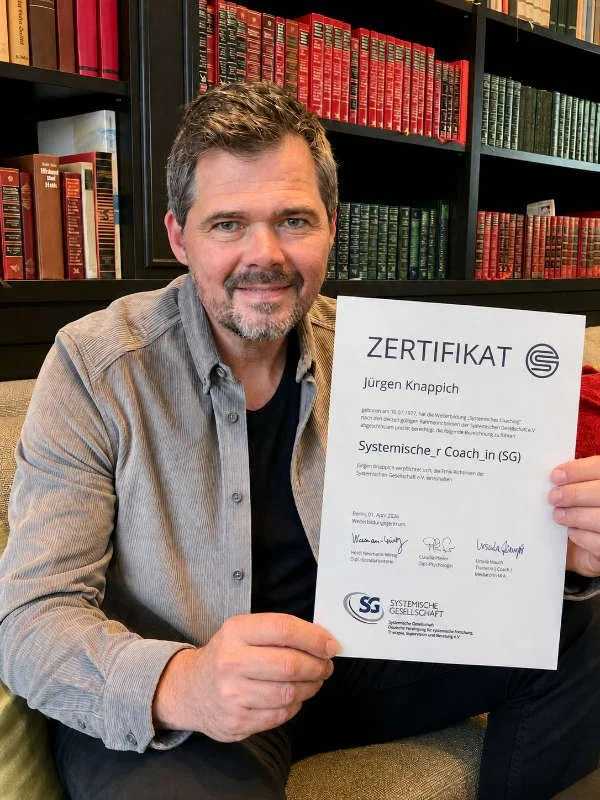 Ein Mann mit grauem Haar und Bart hält ein Zertifikat in den Händen, im Hintergrund sind Bücherregale mit roten und grünen Büchern.