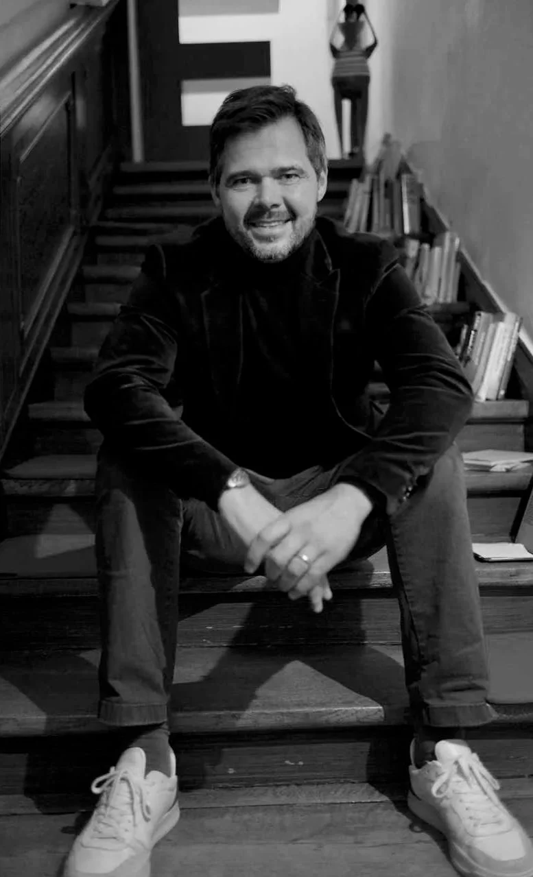 Ein Mann sitzt auf Treppenstufen, trägt einen dunklen Pullover und Sneakers, lächelt und schaut in die Kamera, im Hintergrund sind Bücher und eine Skulptur.