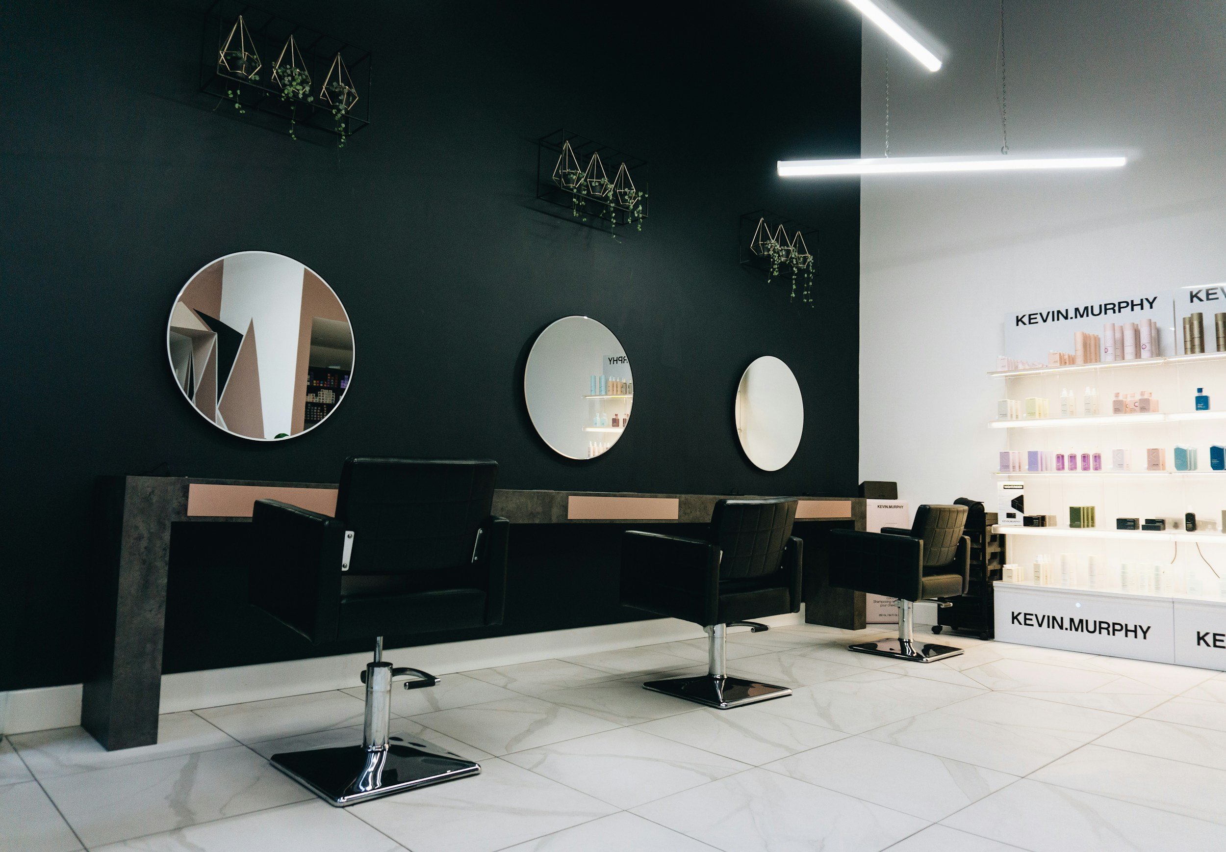 Modern hair salon interior with black chairs, round mirrors, and shelves displaying Kevin Murphy products.