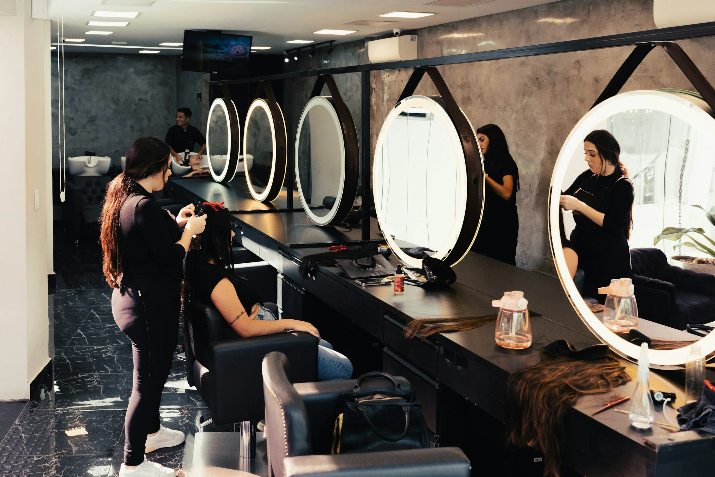 Hair salon with stylists working on clients in front of circular mirrors, tools and hair products on the counter.