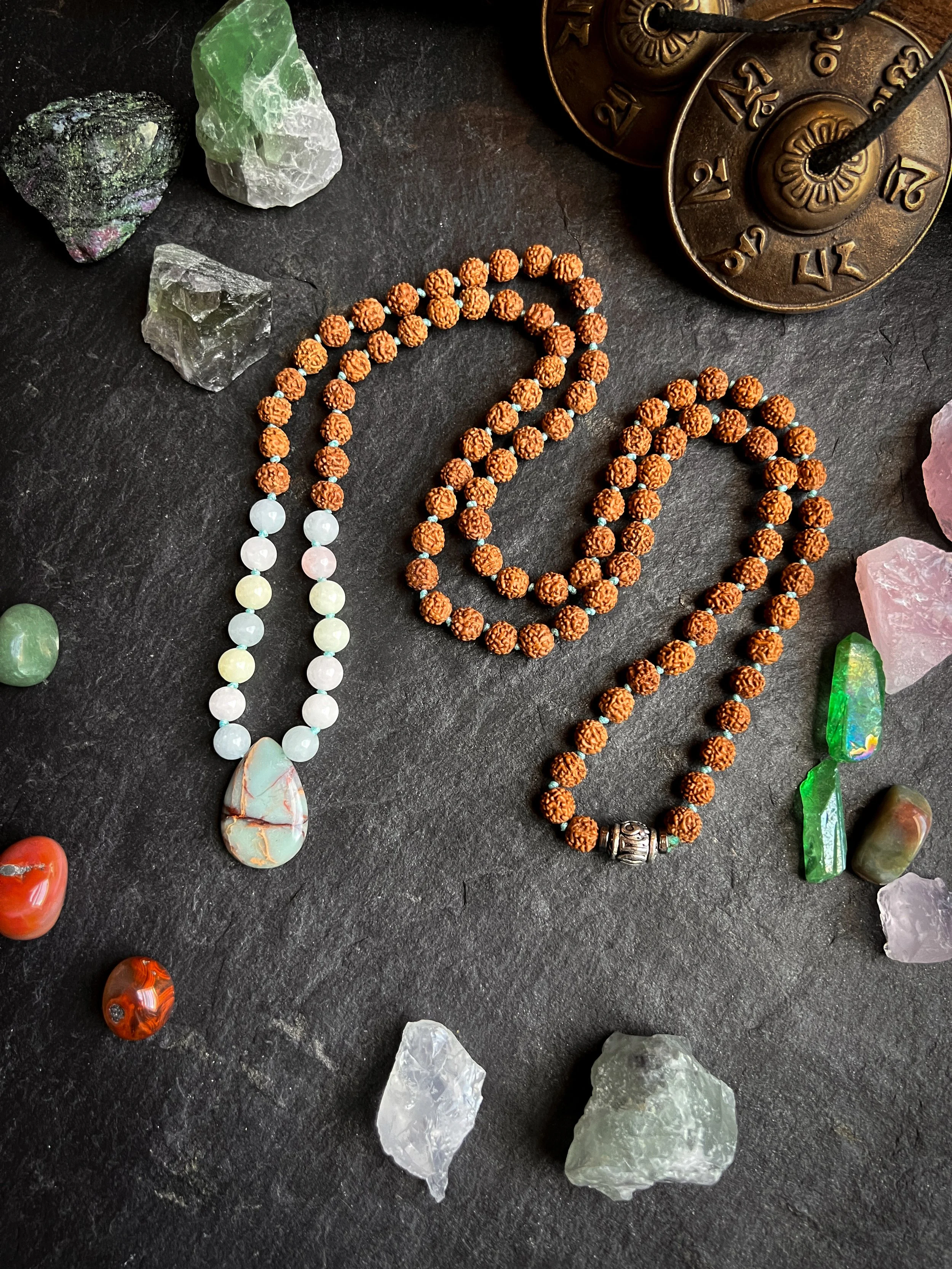 Peaceful arrangement of jewelry and gemstones on a dark surface, including a string of white and pastel beads with a stone pendant, two strands of rudraksha beads, and various colorful crystals and rocks surrounding them.