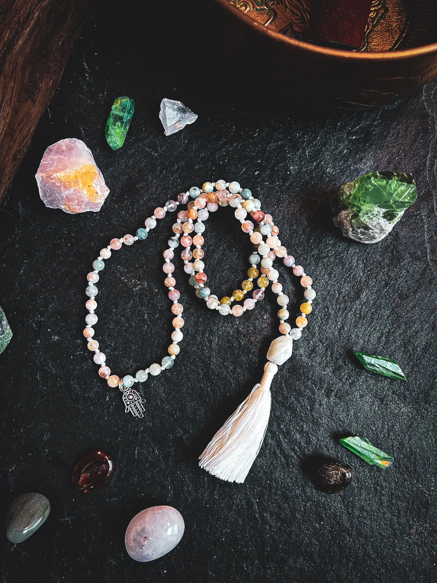 Colorful beaded necklace with white tassel surrounded by various crystals, stones, and a decorative bowl on a black surface.