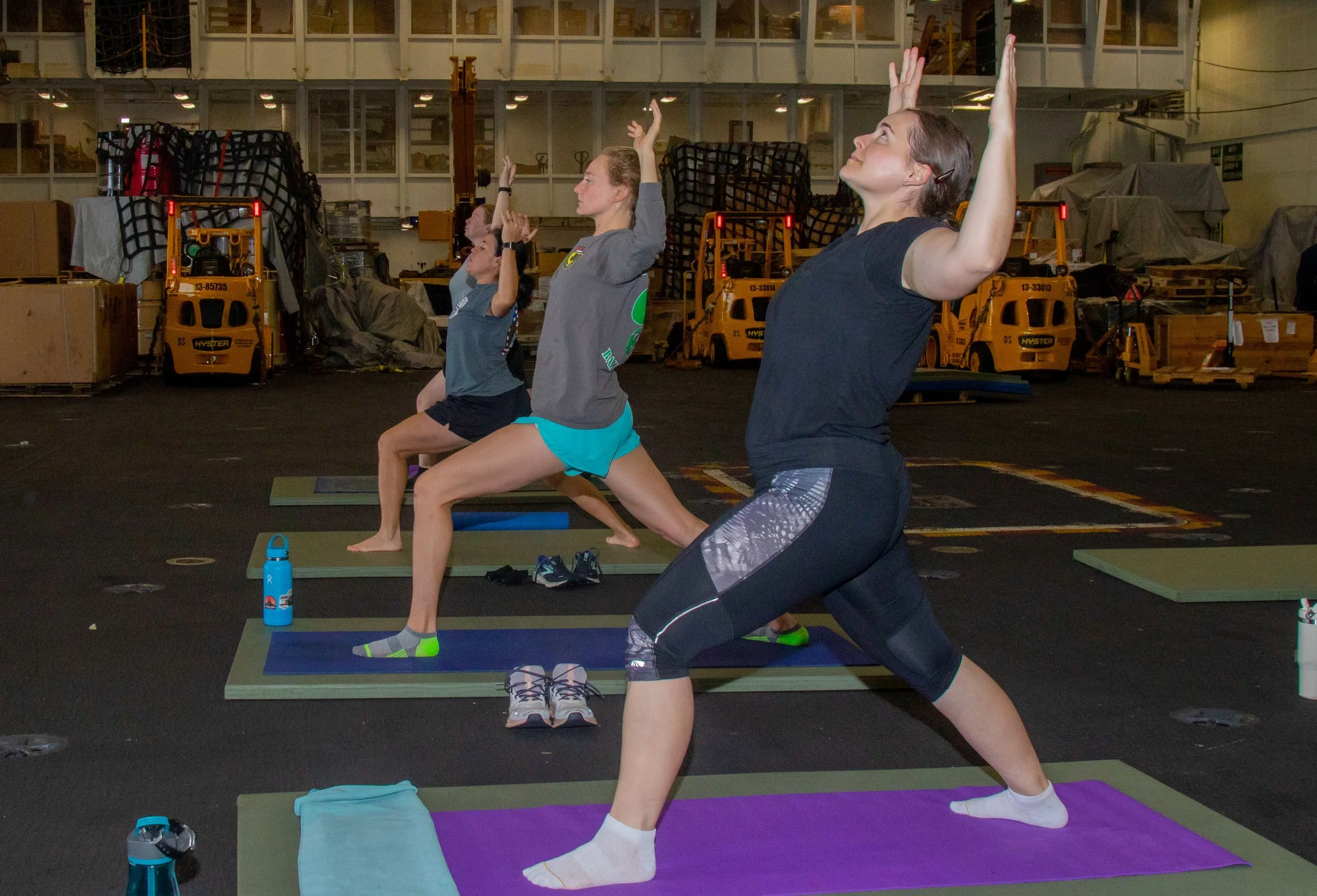 Group of women practicing yoga in a spacious indoor gym with industrial equipment in the background, all in a warrior pose on yoga mats.