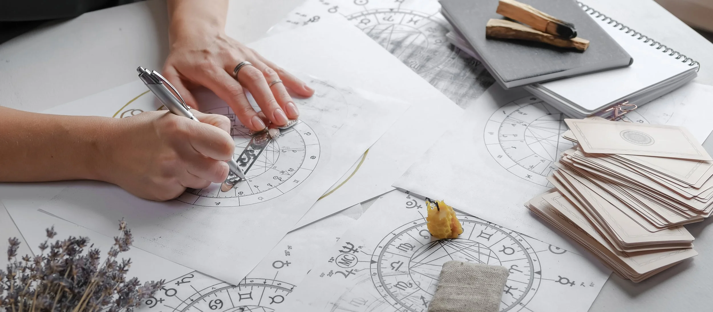 A person is working on astrological charts using a ruler and pen, with various astrology papers, a notebook, and tarot cards spread on the table.