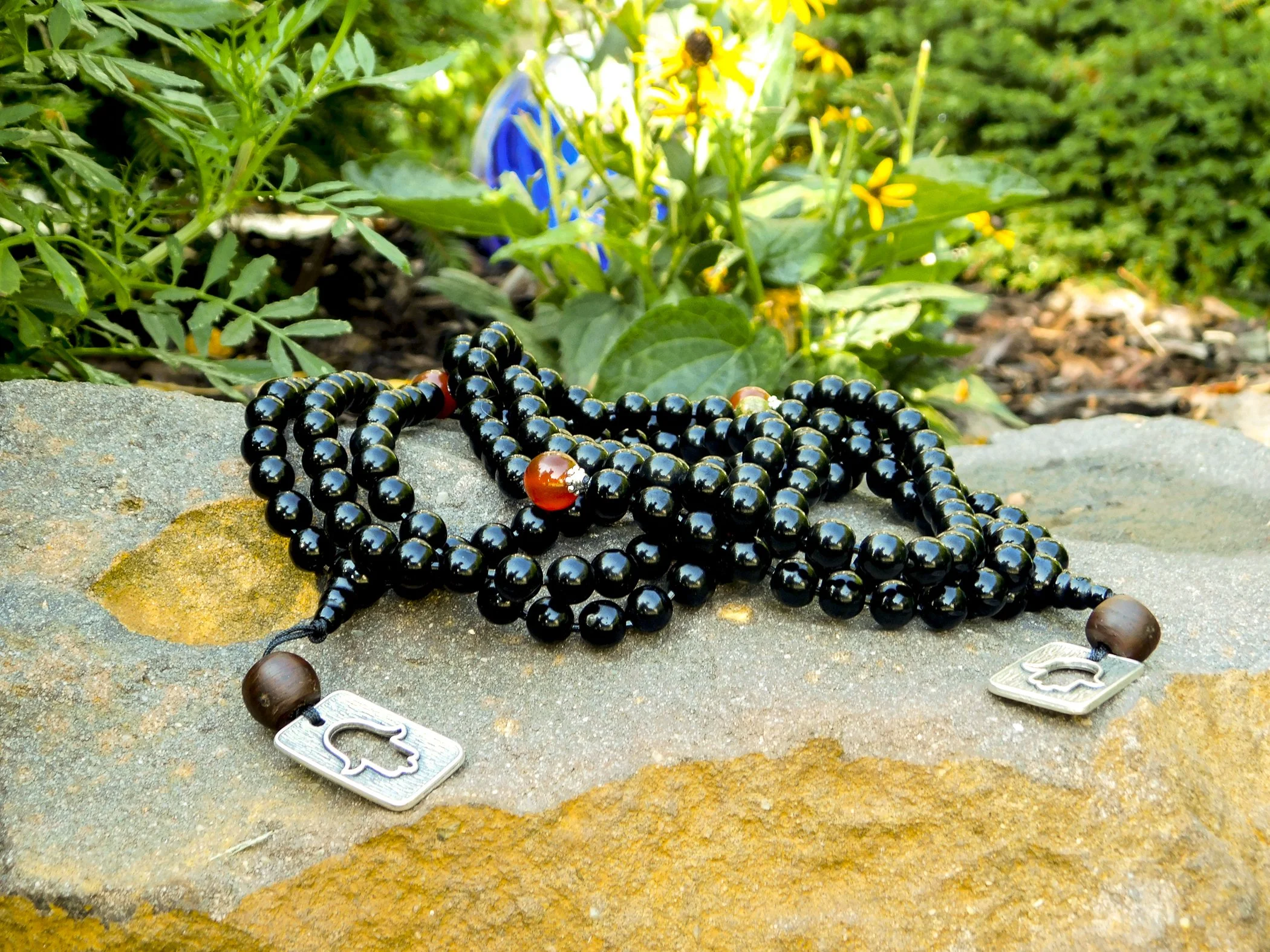 A black beaded necklace with a silver fish charm and a brown bead, resting on a large rock outdoors with green foliage in the background.