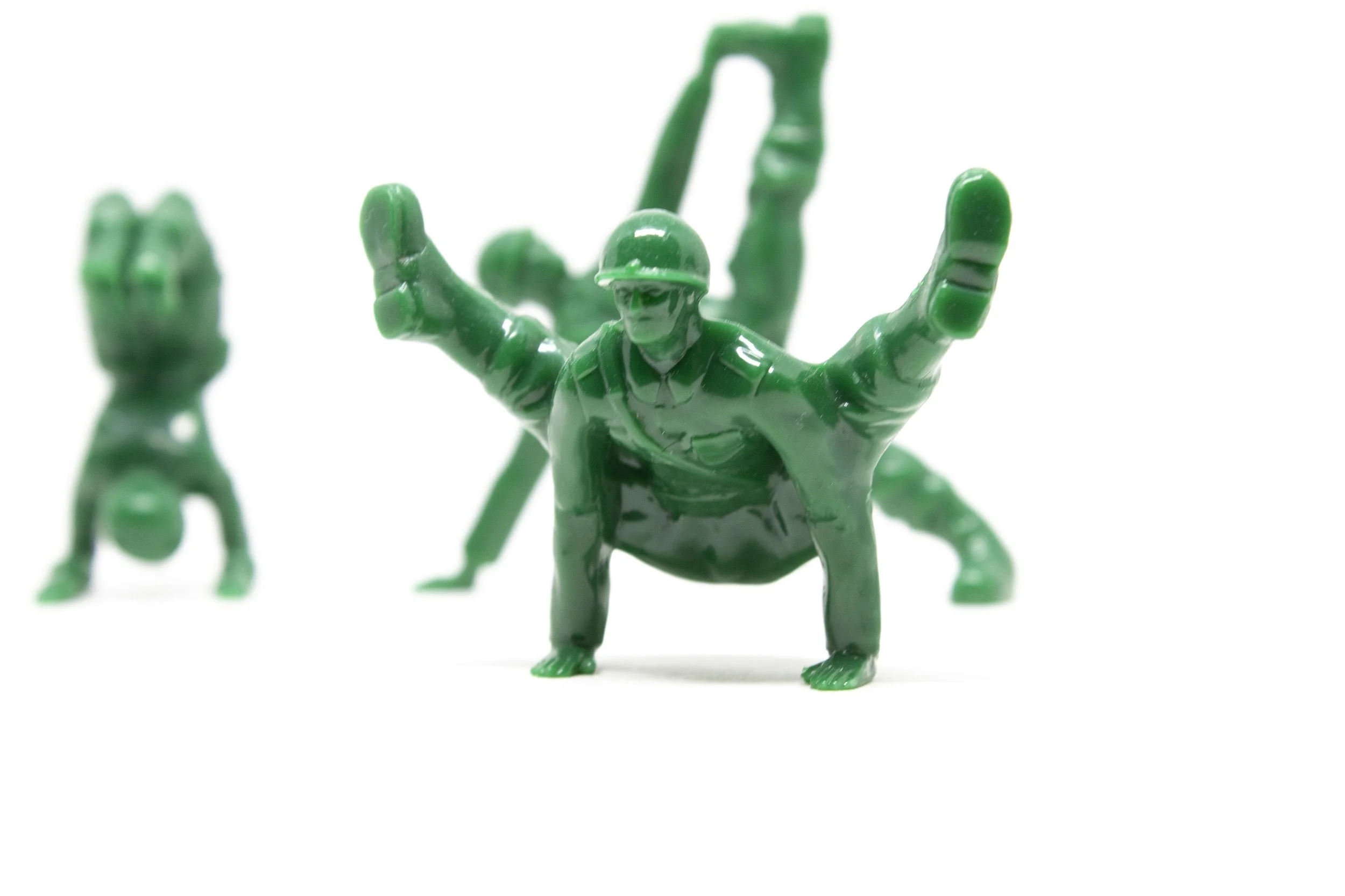 Two green plastic toy soldiers, one doing a handstand and the other in a push-up position, with a third soldier in the background holding a rifle.