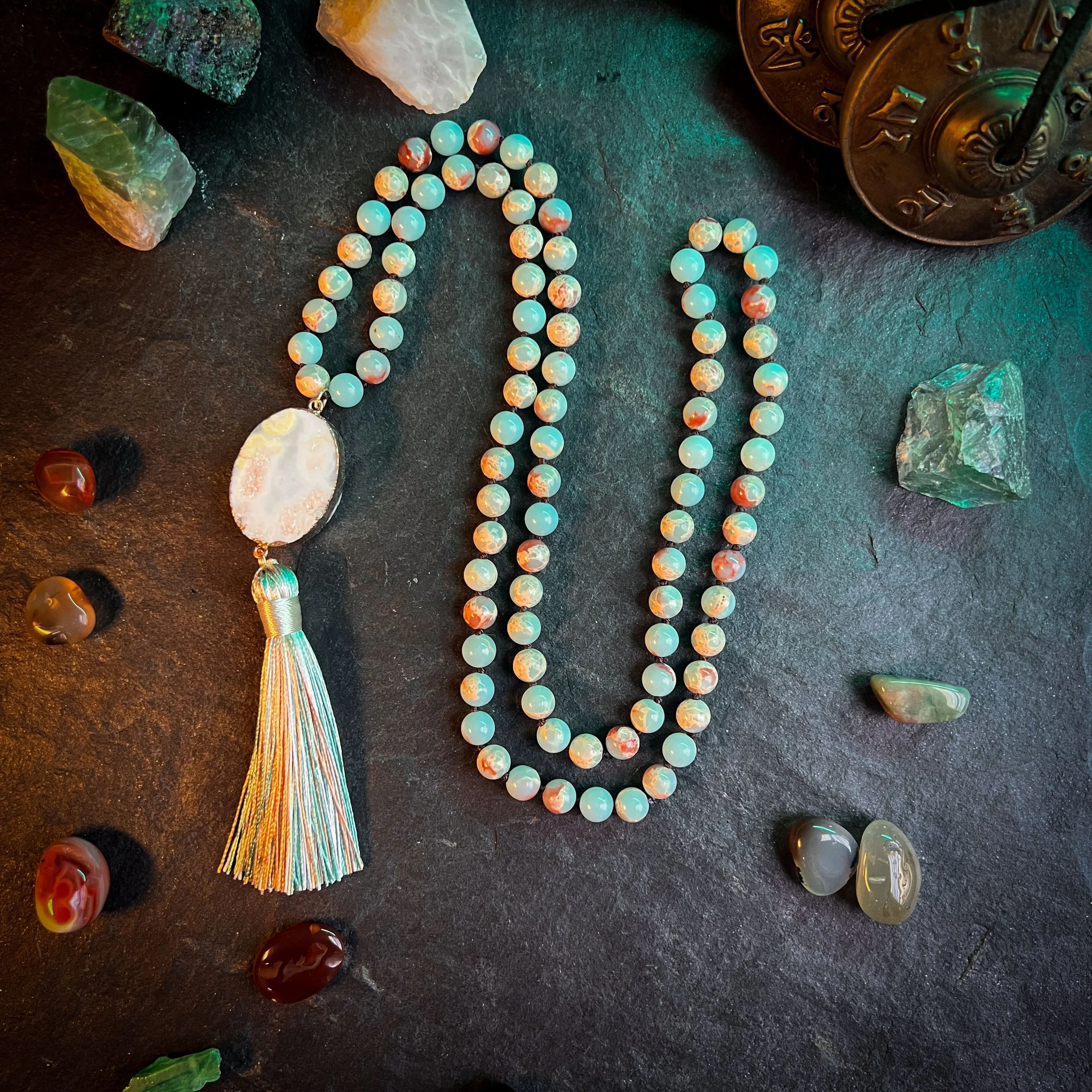 A beaded necklace with a large oval pendant and colorful tassel lies on a dark surface, surrounded by various gemstones and decorative objects.
