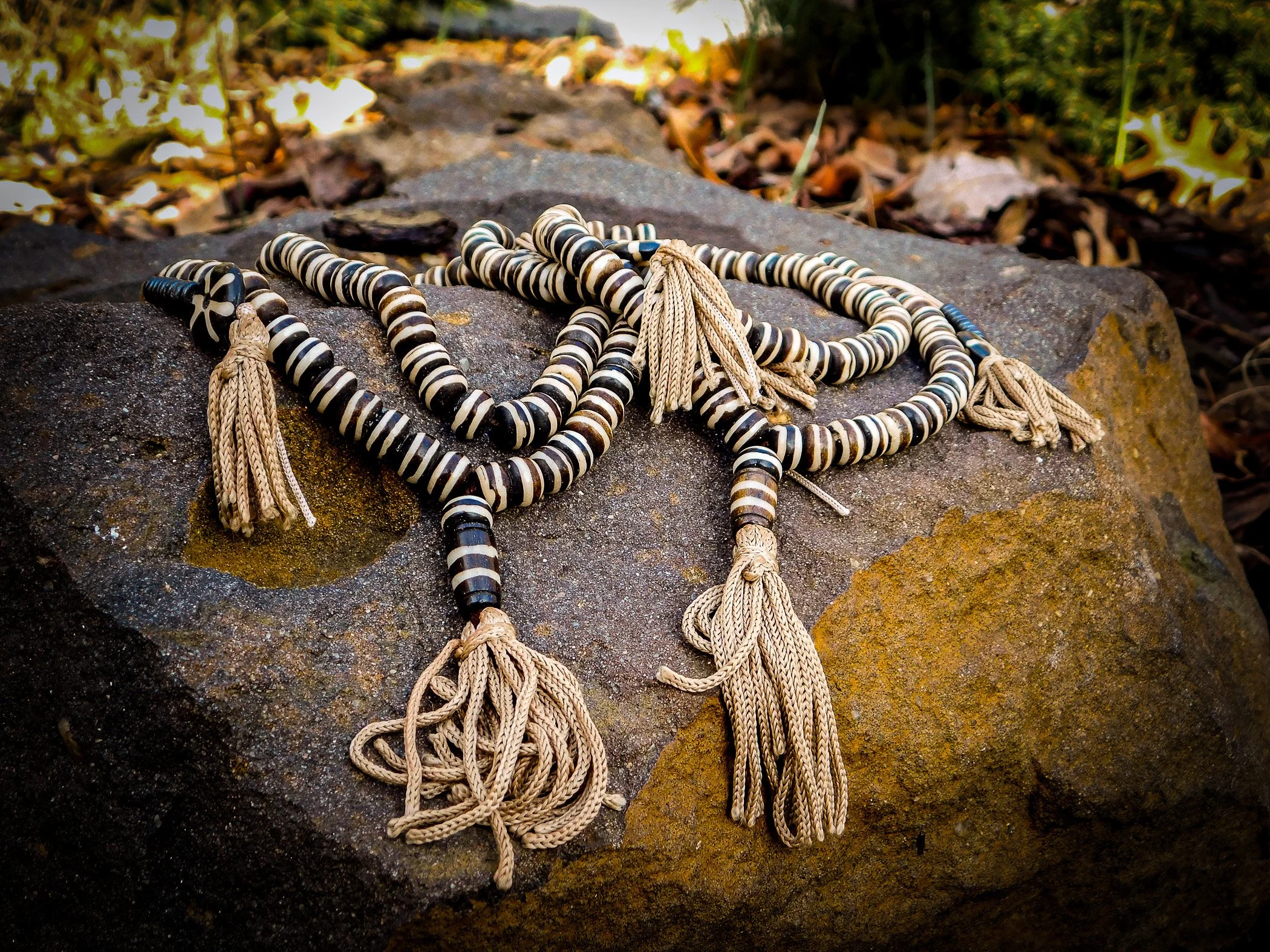 Three beaded necklaces with black and white stripes and beige tassels lying on a large gray rock, surrounded by fallen leaves and natural outdoor scenery.