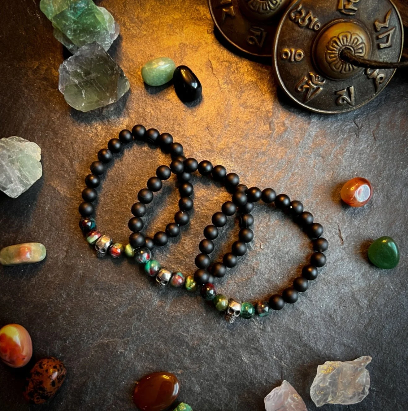 Two black beaded necklaces with multicolored beads and silver skull beads lying on a dark textured surface surrounded by various semi-precious stones and crystals. A vintage metal device with numbered dials is partially visible in the upper right corner.