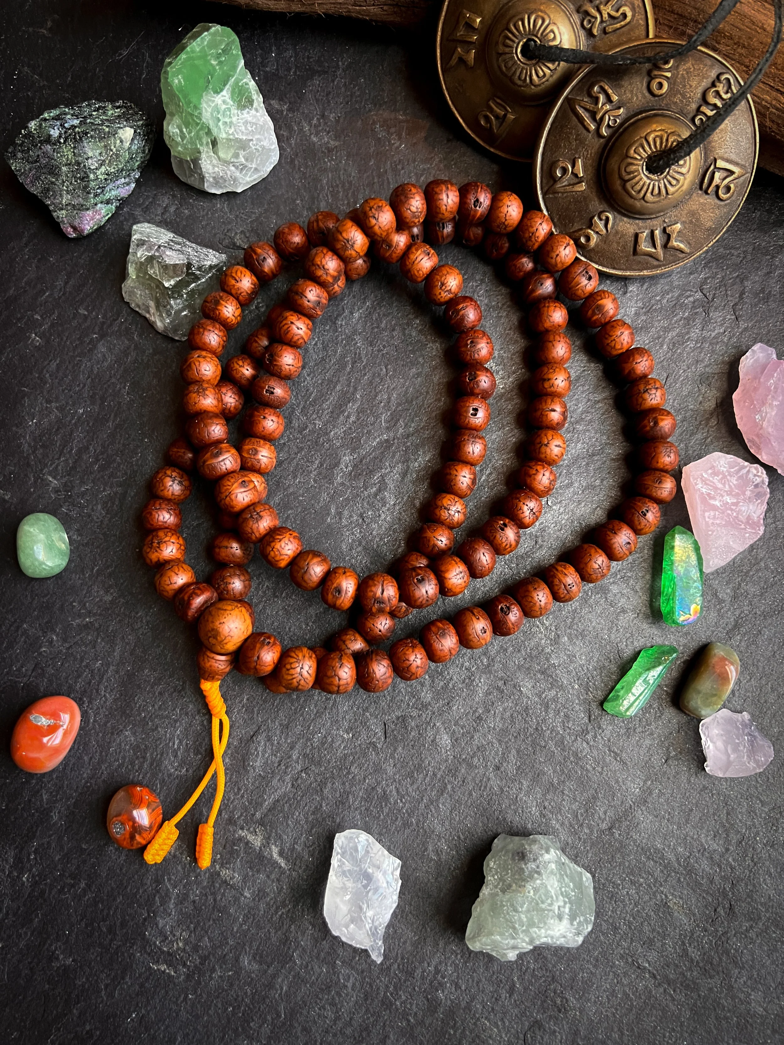Brown beaded mala necklace with a yellow tie, surrounded by various colorful gemstones on a dark surface, with brass prayer wheels in the background.