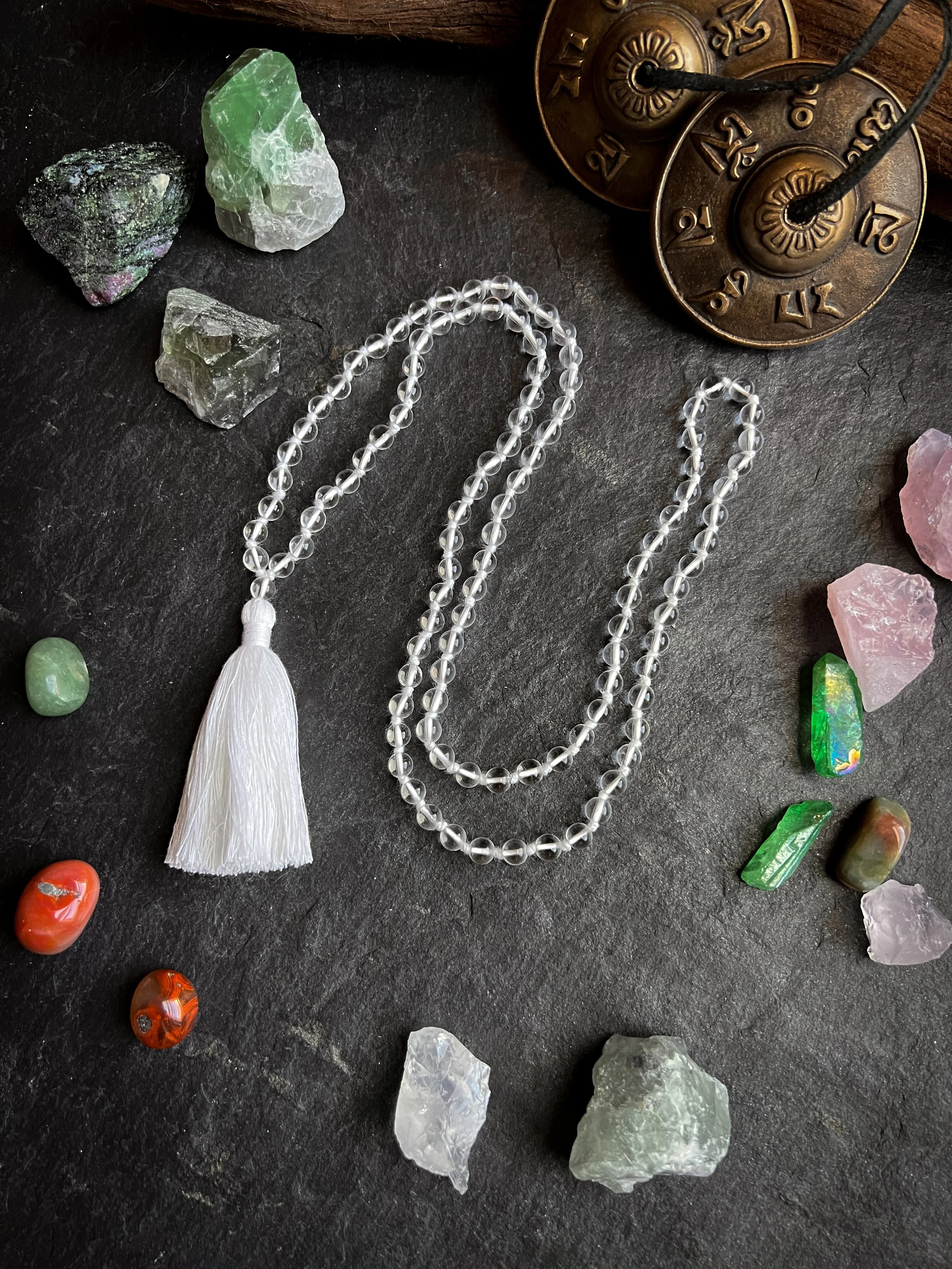 A string of clear beads with a white tassel, surrounded by various colorful gemstones, and vintage clock parts on a dark surface.