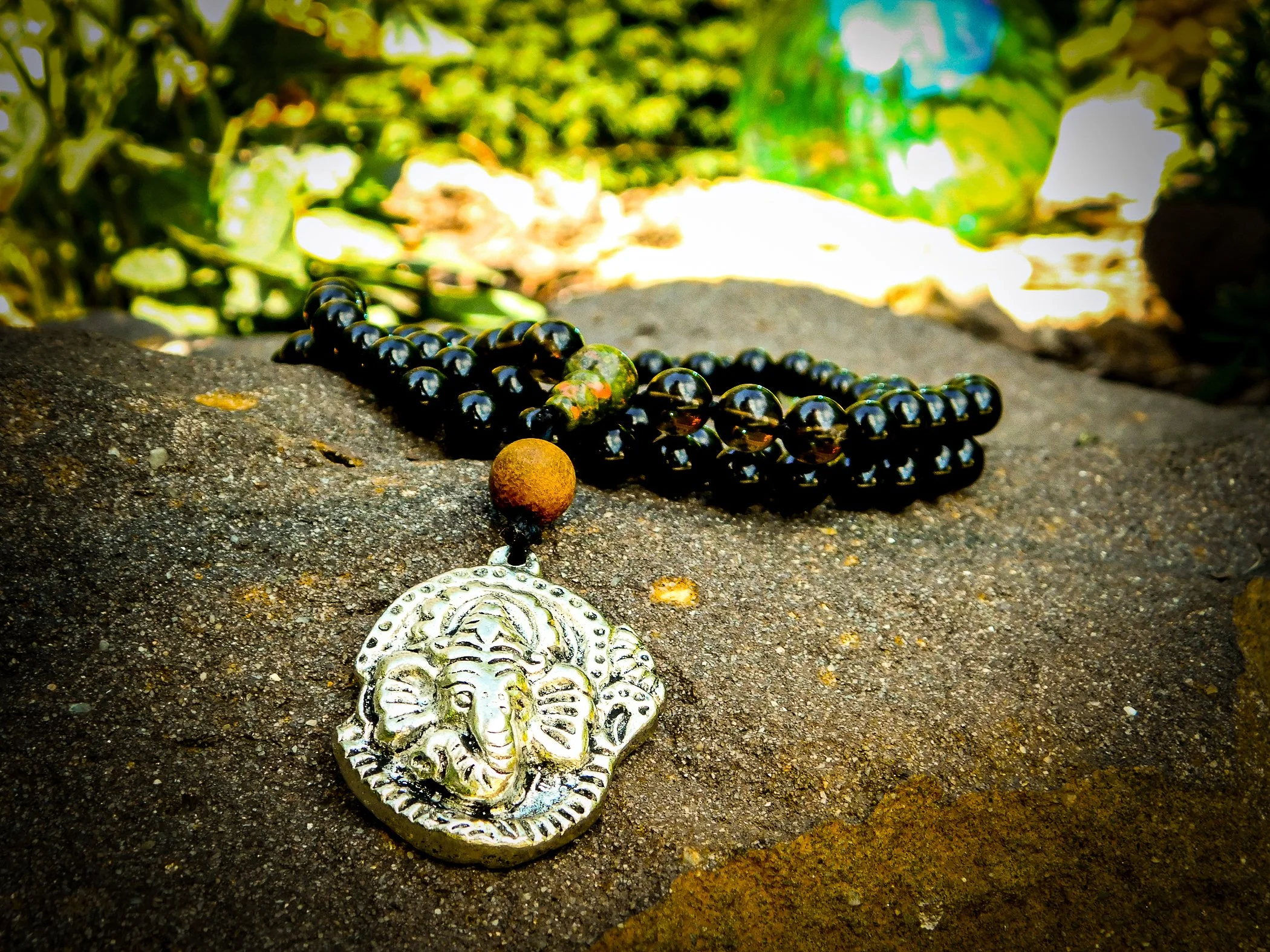 A silver pendant with an elephant design, a rust-colored bead, and black beaded jewelry with an iridescent bead, resting on a rock outdoors.