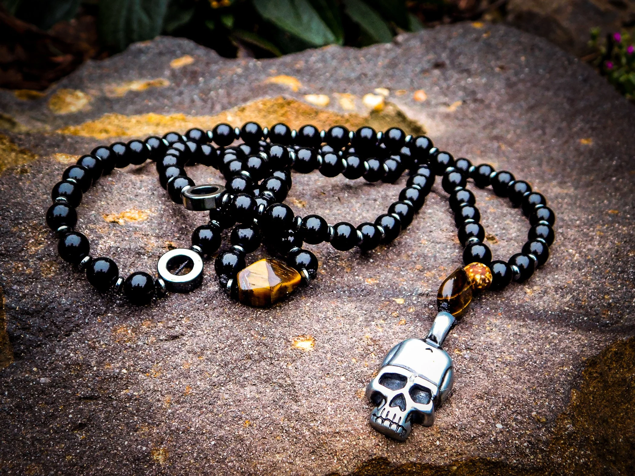 Black bead necklace with skull pendant and amber-colored beads, resting on a textured rock surface.
