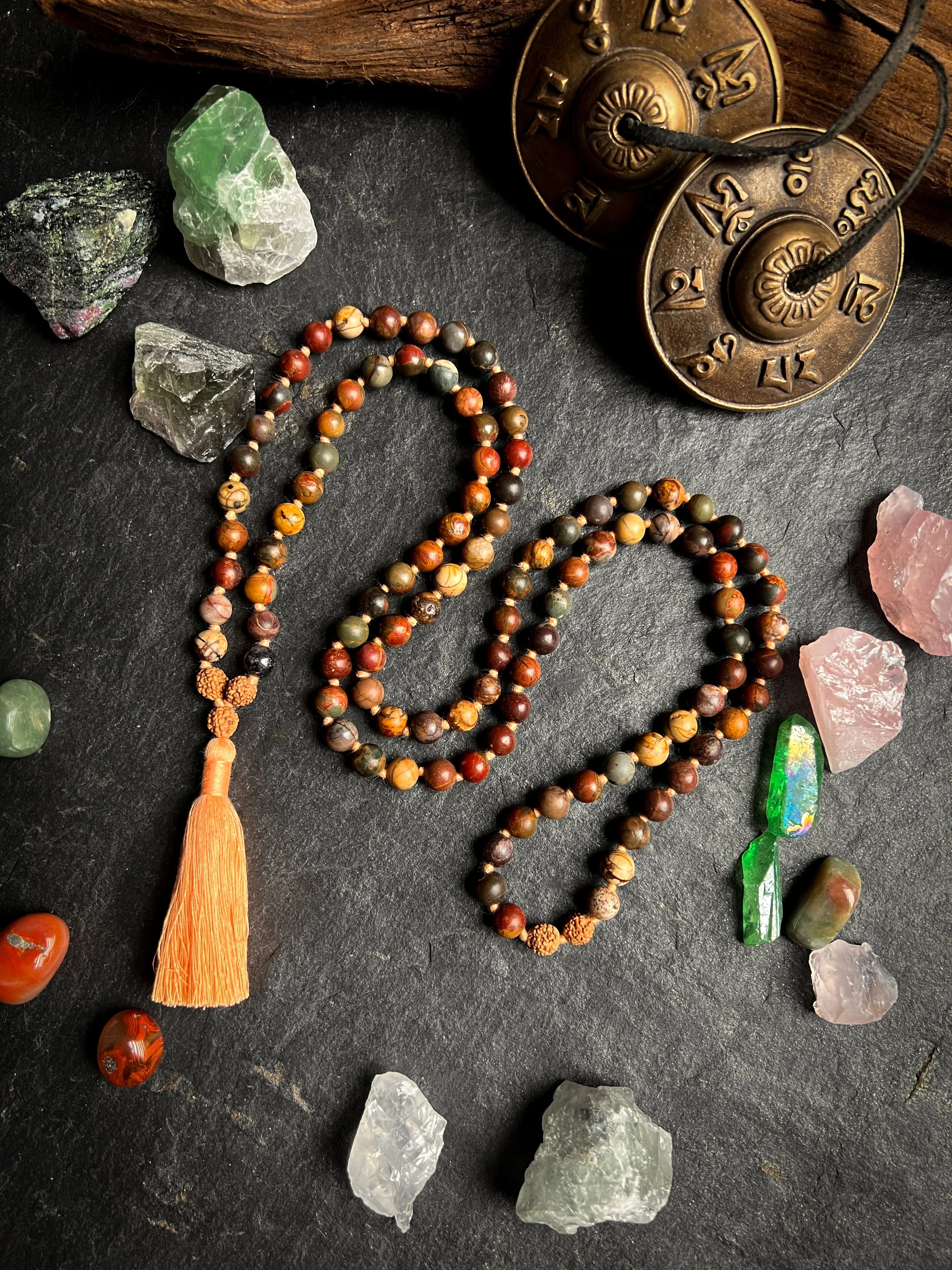 A close-up of a collection of various gemstones and crystals on a black surface, with some beaded jewelry and a vintage brass clock in the background.
