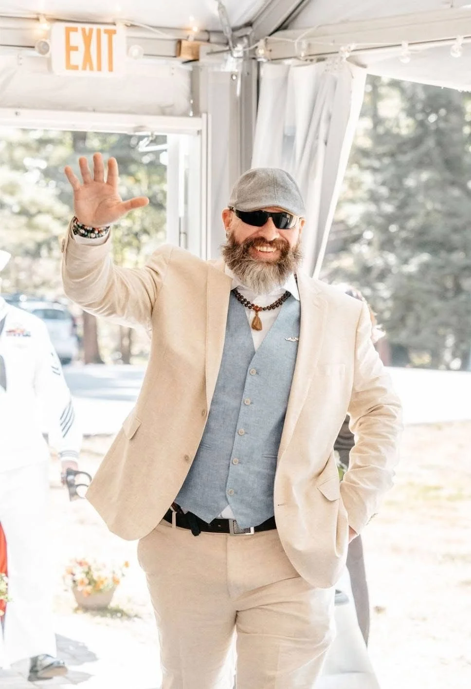Smiling man with a beard, sunglasses, and a gray flat cap waving, dressed in a beige suit and light blue vest, inside a white tent with an 'EXIT' sign overhead.