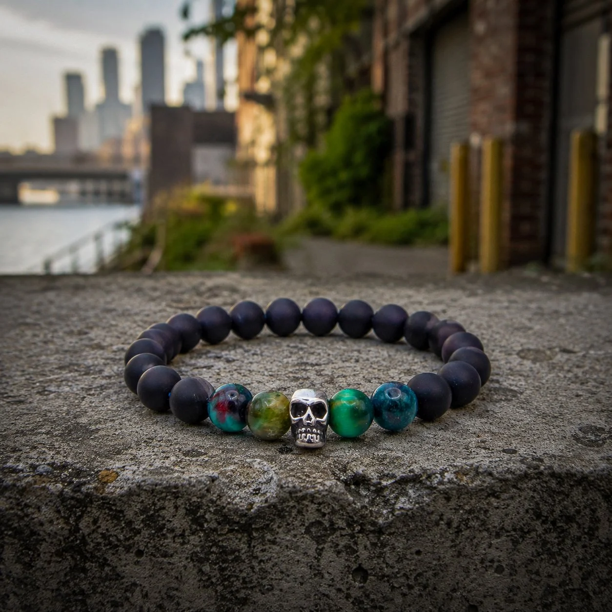 A beaded bracelet with black beads and a silver skull bead, placed on a concrete surface outdoors with a city skyline and river in the background.