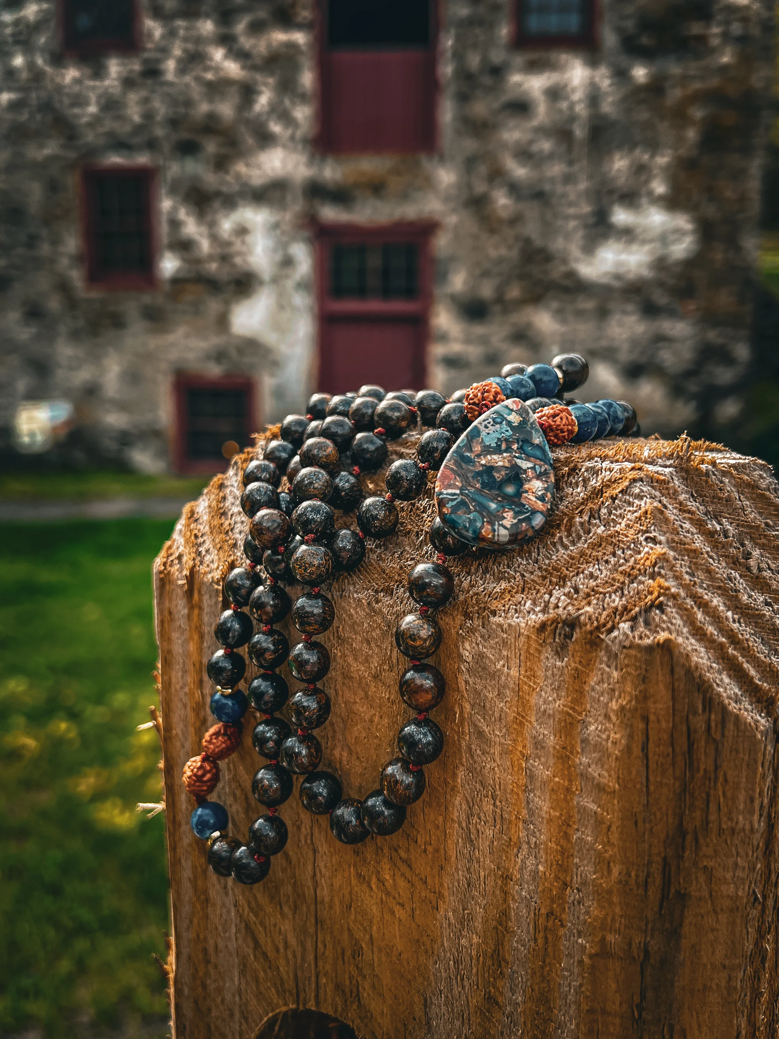 A beaded necklace with black and brown beads, and a large decorative pendant, draped over a piece of wood outside with an old stone building with small windows in the background.