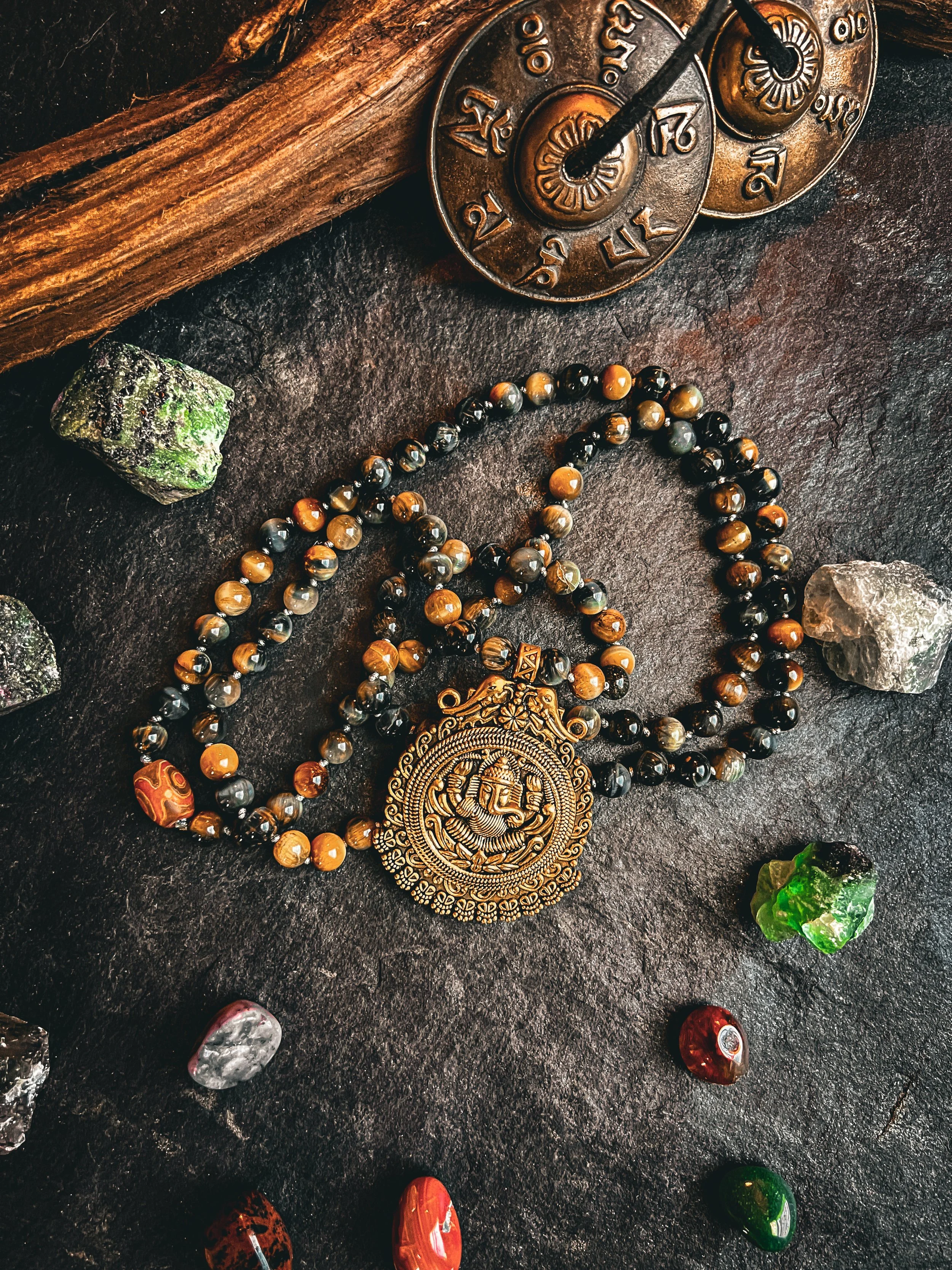 A beaded necklace with a gold medallion featuring Lord Ganesha, surrounded by colorful semi-precious stones, a wooden stick, and two metallic circular objects with numerals and symbols, all placed on a dark textured surface.