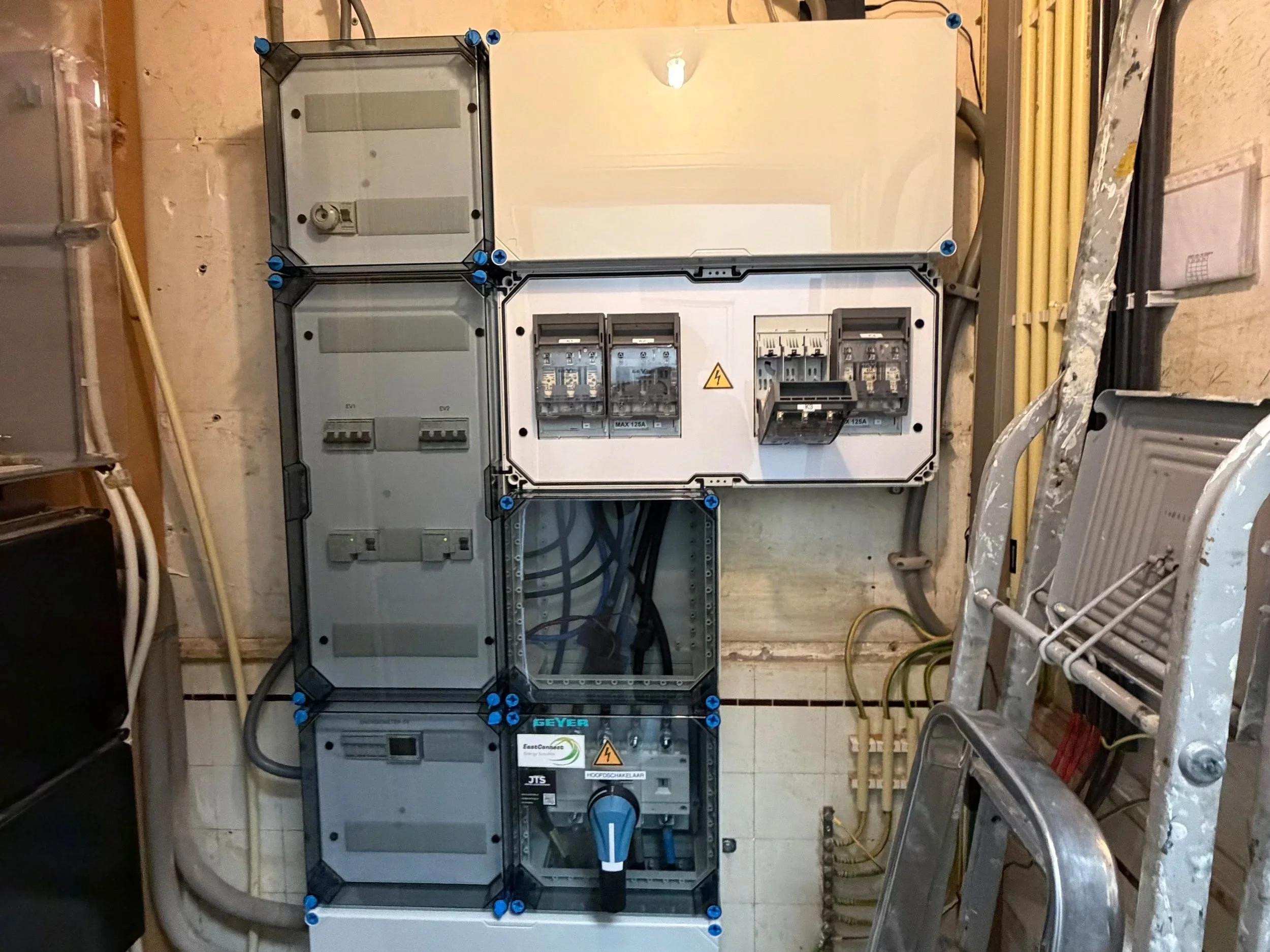 Binnenin een elektrisch verdeelbord met verschillende schakelaars en kabels, op een wand met enkele leidingen en een ladder ernaast.