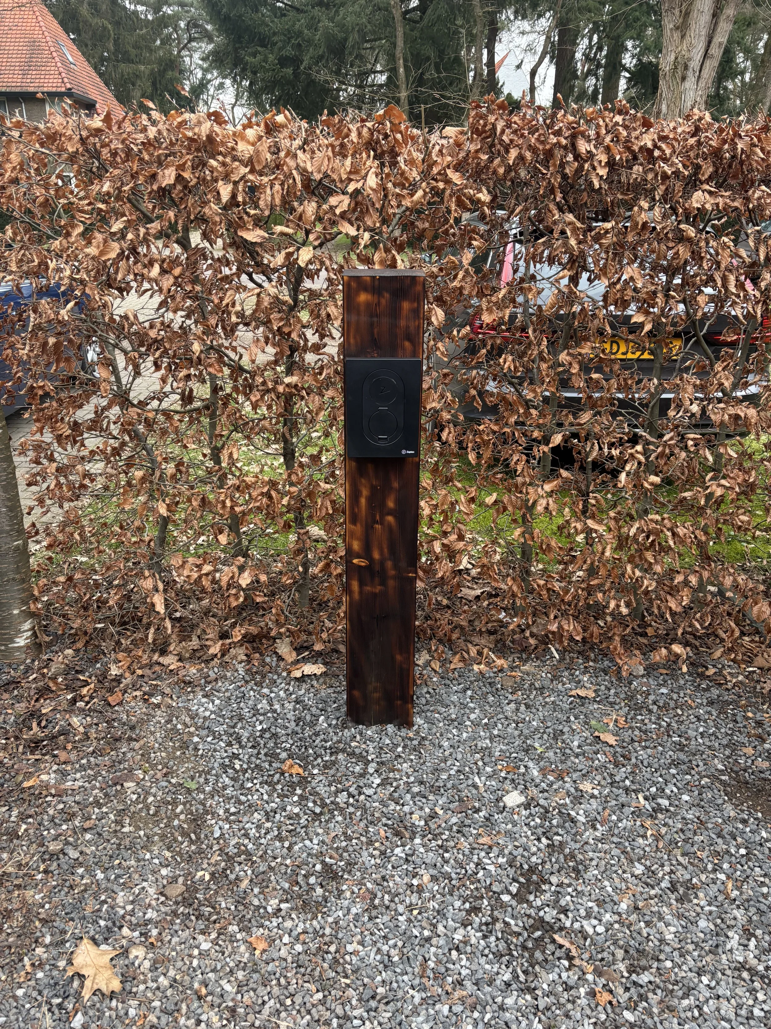 Een houten paal met een black box elektrische deurbel onder een boom met verwelkte bladeren.