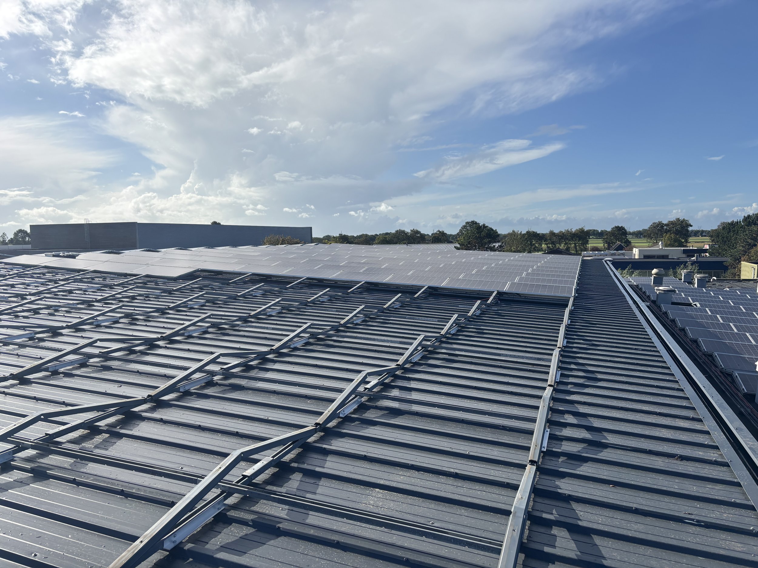 Een plat dak met metalen bedekking en zonnepanelen onder een deels bewolkte blauwe hemel.