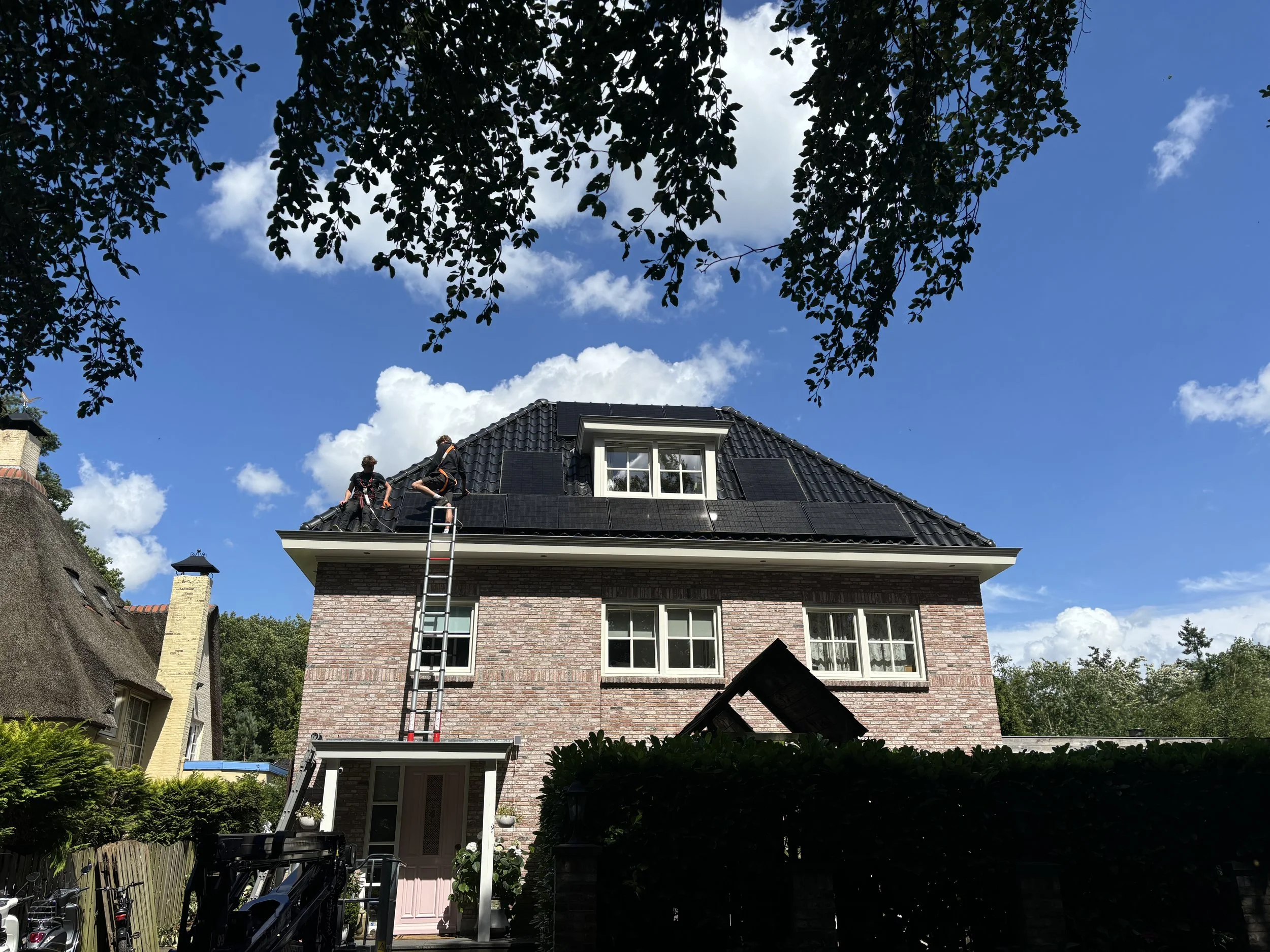 Twee mensen werken op het dak van een huis met een ladder, onder een blauwe hemel met wat wolken.