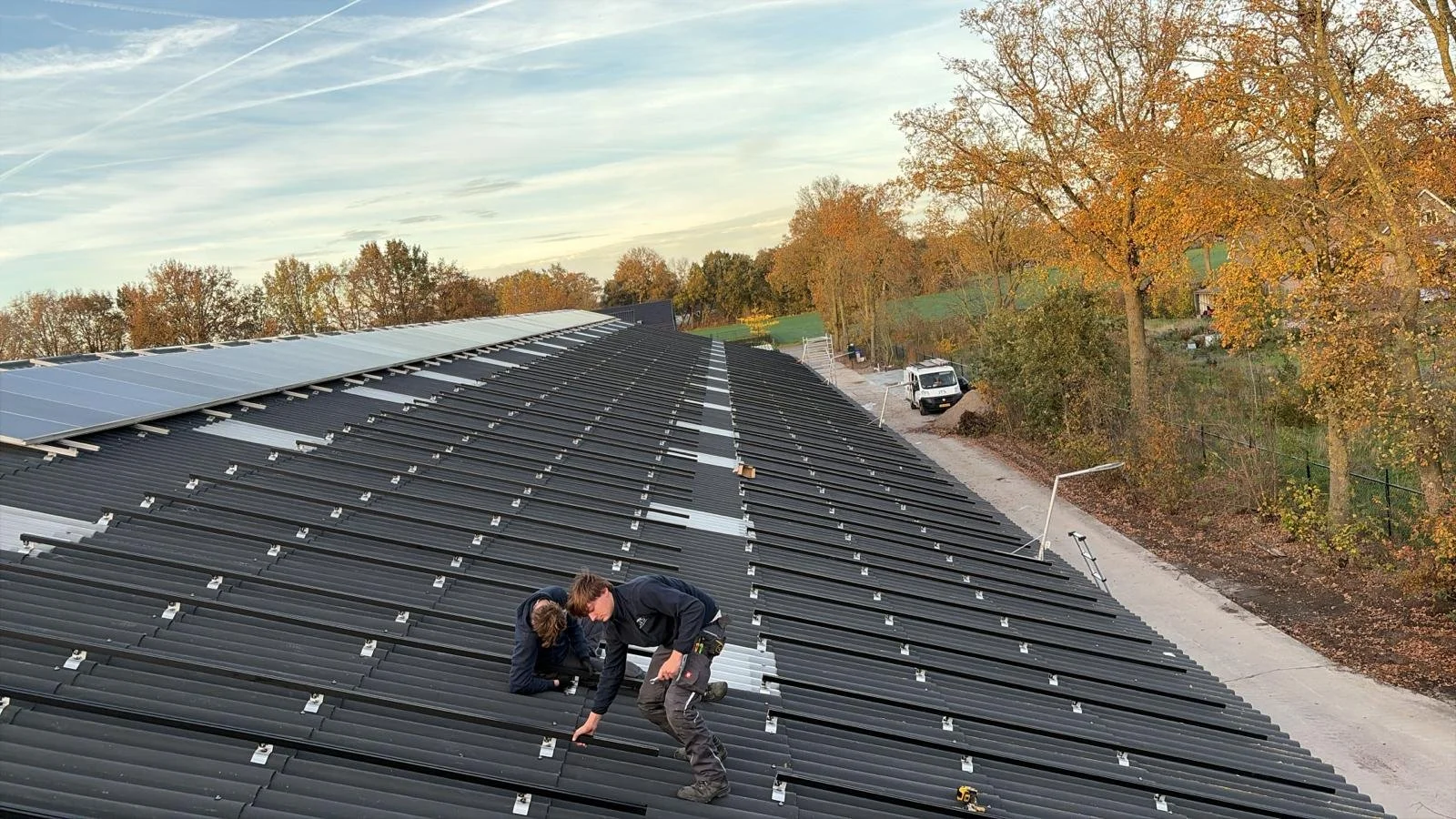 Twee mensen installeren zonnepanelen op een zwart metalen dak, omringd door bomen met herfstkleuren.