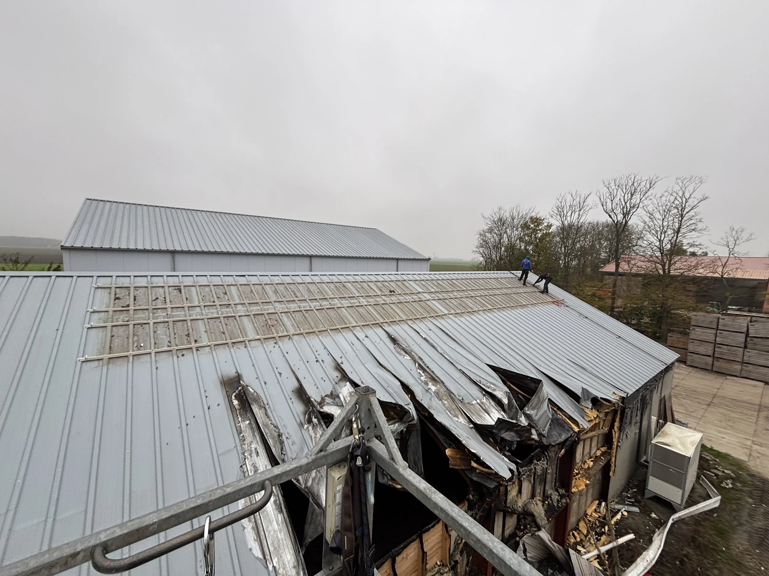 Verschillende mannen werken op het metalen dak van een gestoord gebouw met schade veroorzaakt door brand, met op de achtergrond een industrieel landschap.
