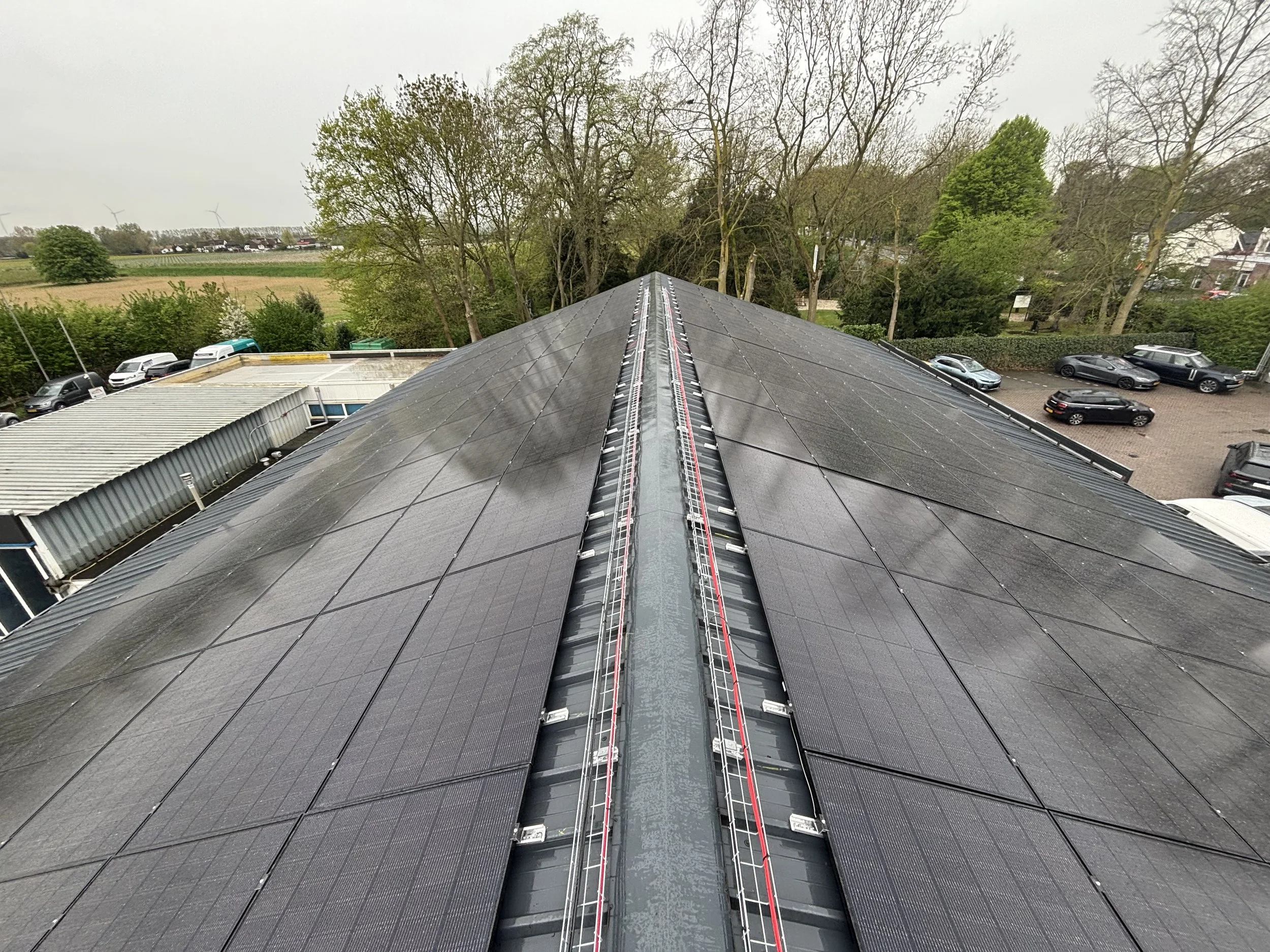 Zonnedak met zonnepanelen, enkele panelen en het dakbedekkingsmateriaal zien er nat uit. Er staan bomen en een parkeerplaats met auto's op de achtergrond.