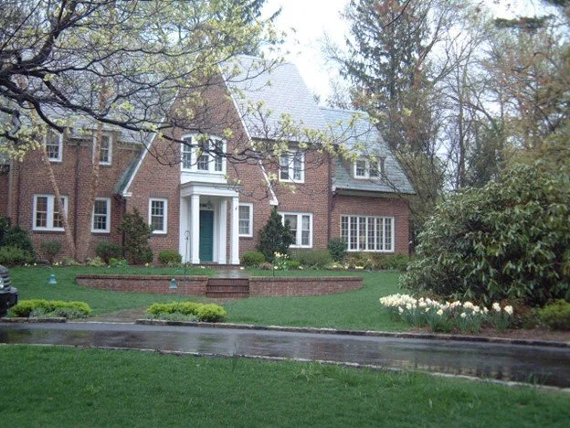 A large brick house with a steep gray roof, white trim, and multiple windows, surrounded by a lush green lawn and garden beds, with trees and clouds in the sky.