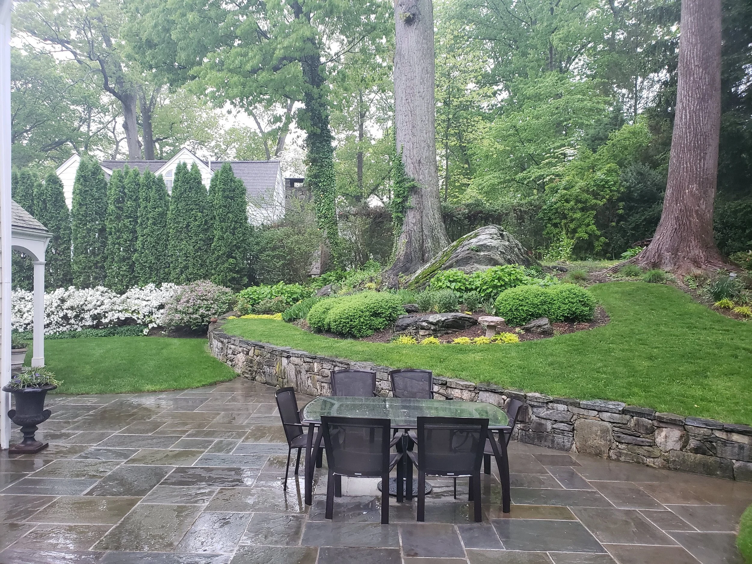 Rain-soaked patio with black patio table and chairs, stone retaining wall, lush green garden with large trees, bushes, and flowering plants.