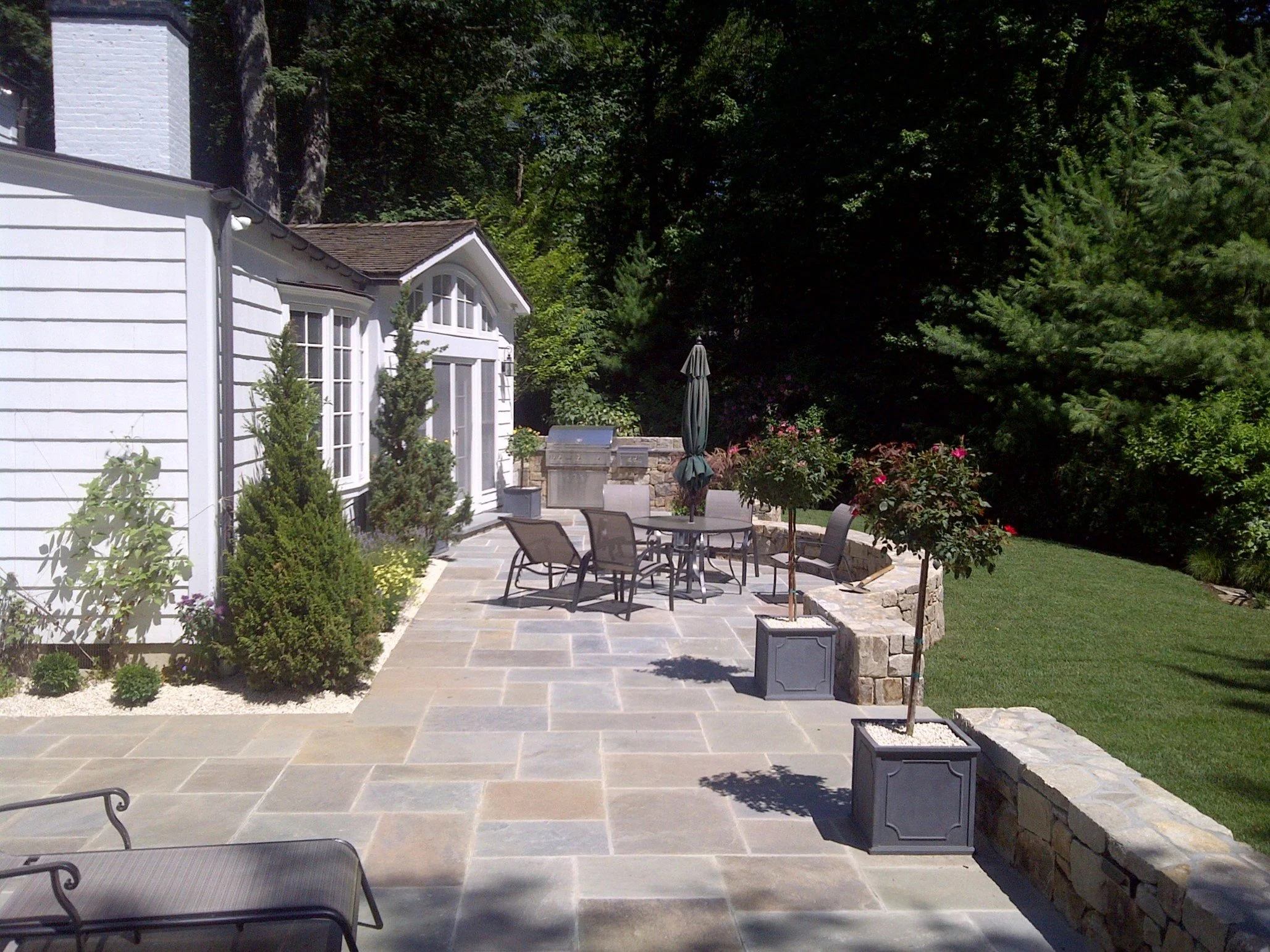 A backyard patio with a table and chairs, potted trees, a barbecue grill, and surrounded by greenery and a stone wall.