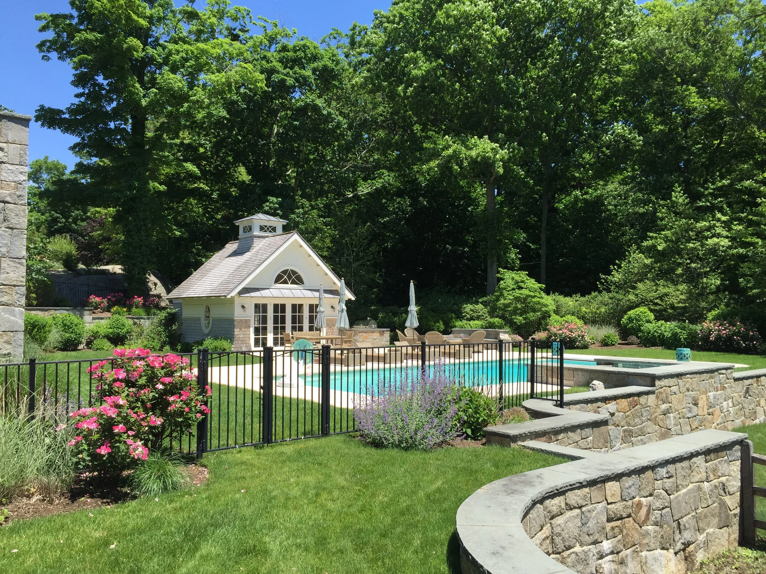 A backyard with a swimming pool, surrounded by a stone wall and black metal fence, with a small white pool house and lush green trees in the background.