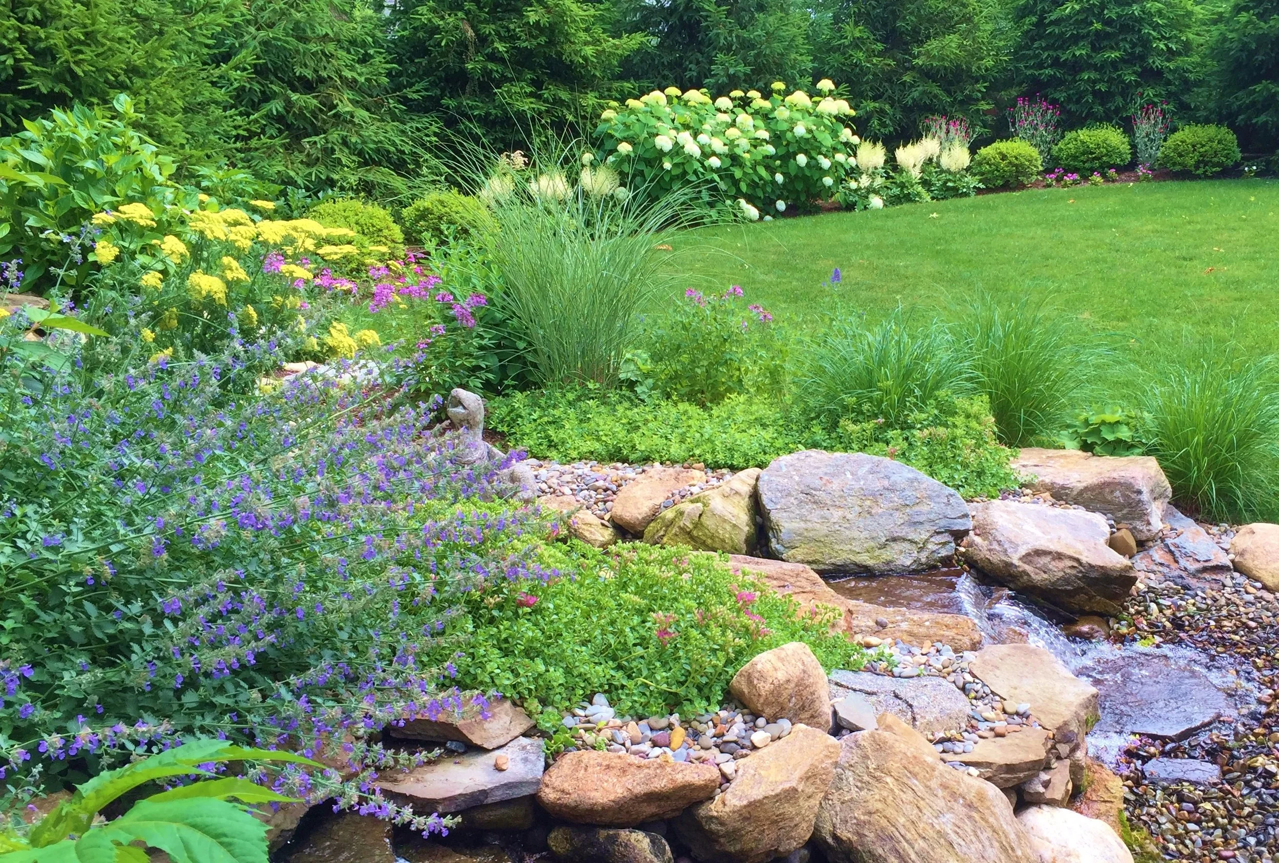 A lush garden with various colorful flowers, green bushes, and a small rocky stream flowing through it.