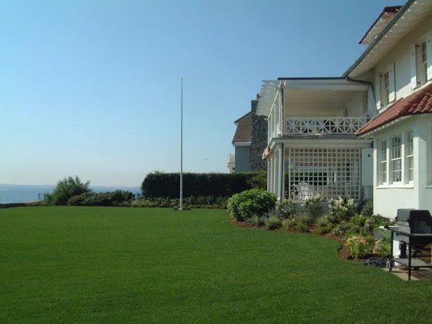 Lawn with a house featuring a porch and a balcony, overlooking the ocean in the background.