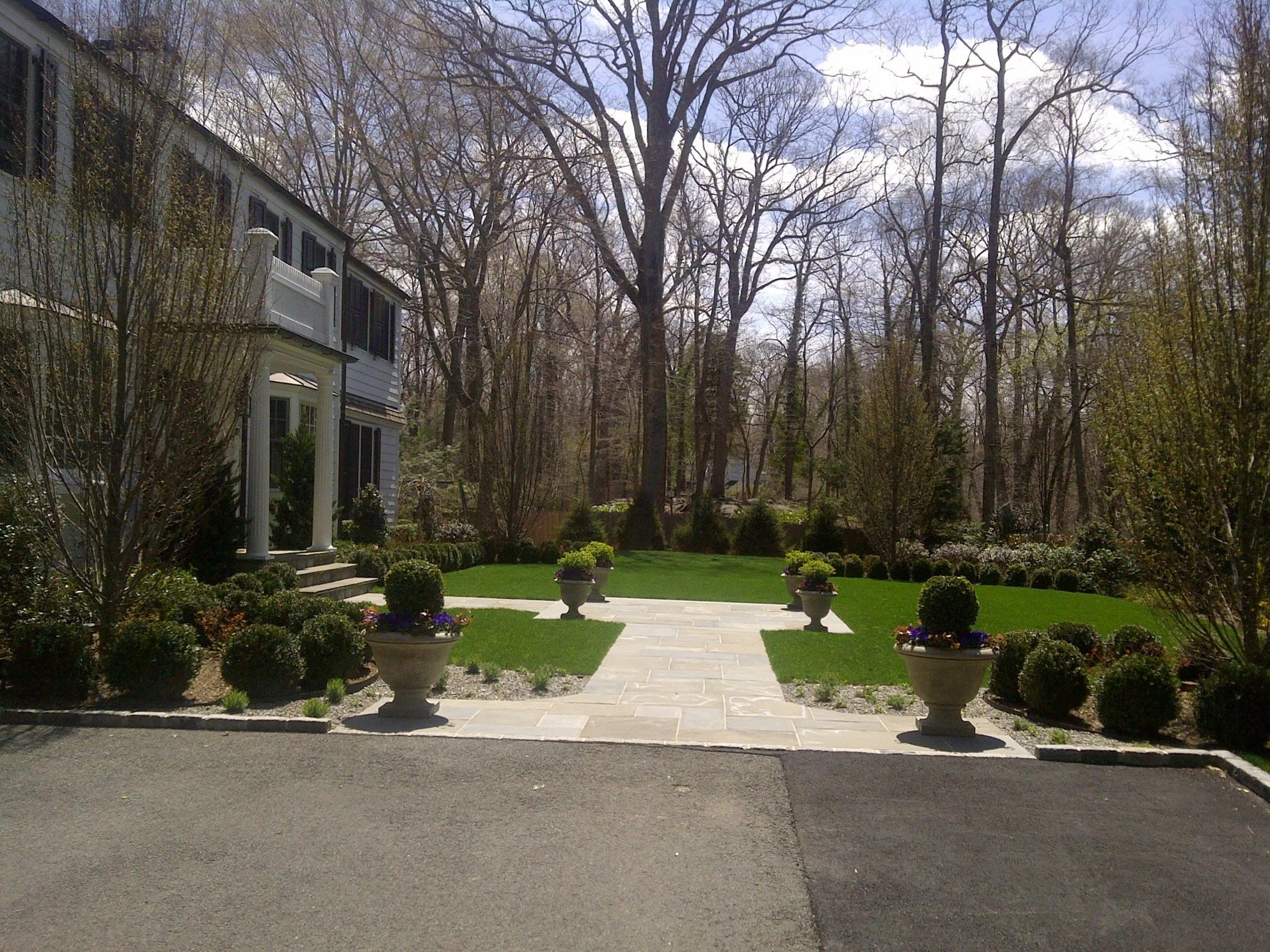 A well-maintained backyard garden with green grass, potted plants, and a paved walkway leading to a wooded area, next to a white two-story house with black shutters.