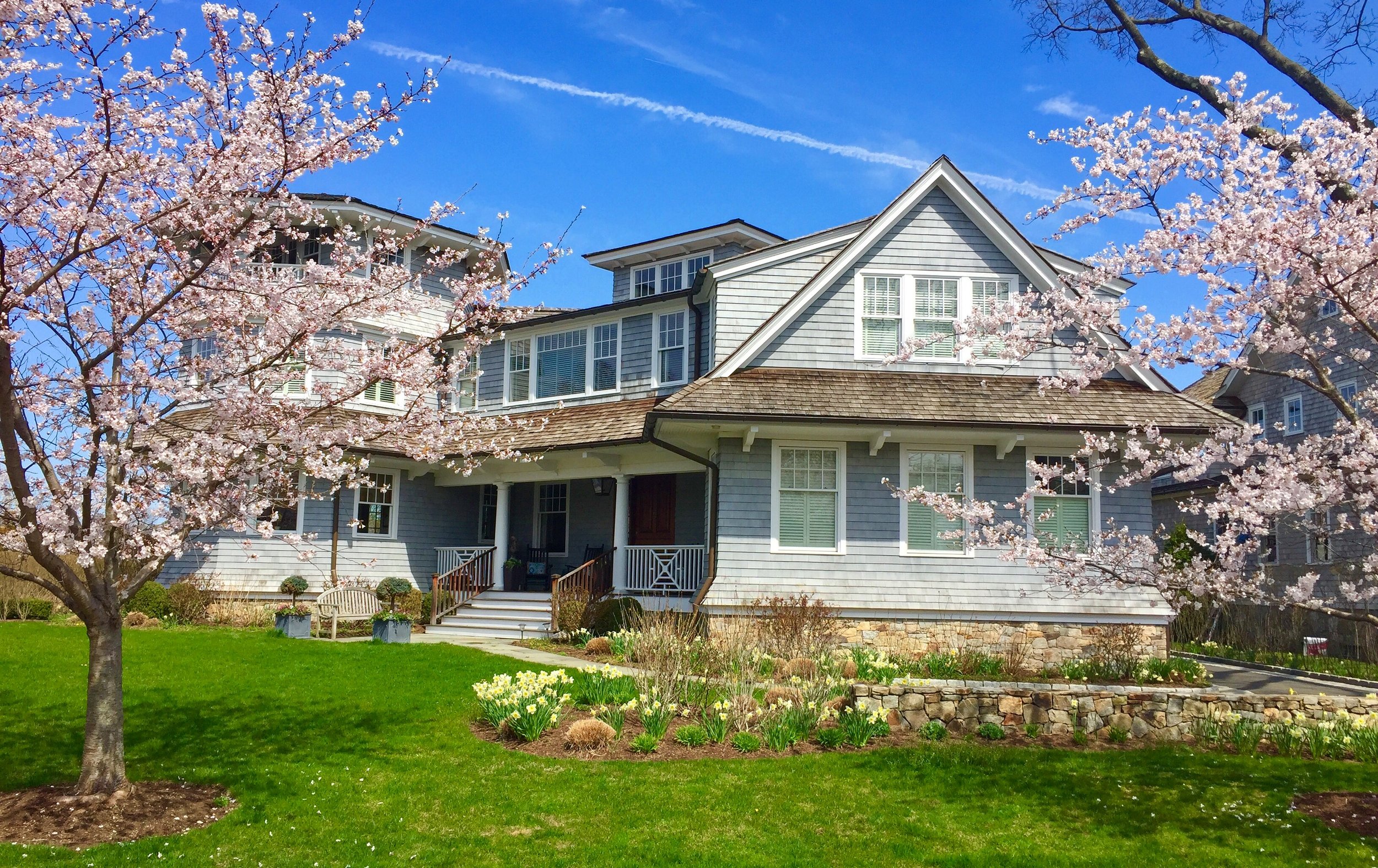 A light blue, multi-story house with a landscaped yard featuring blooming trees and spring flowers, under a blue sky.