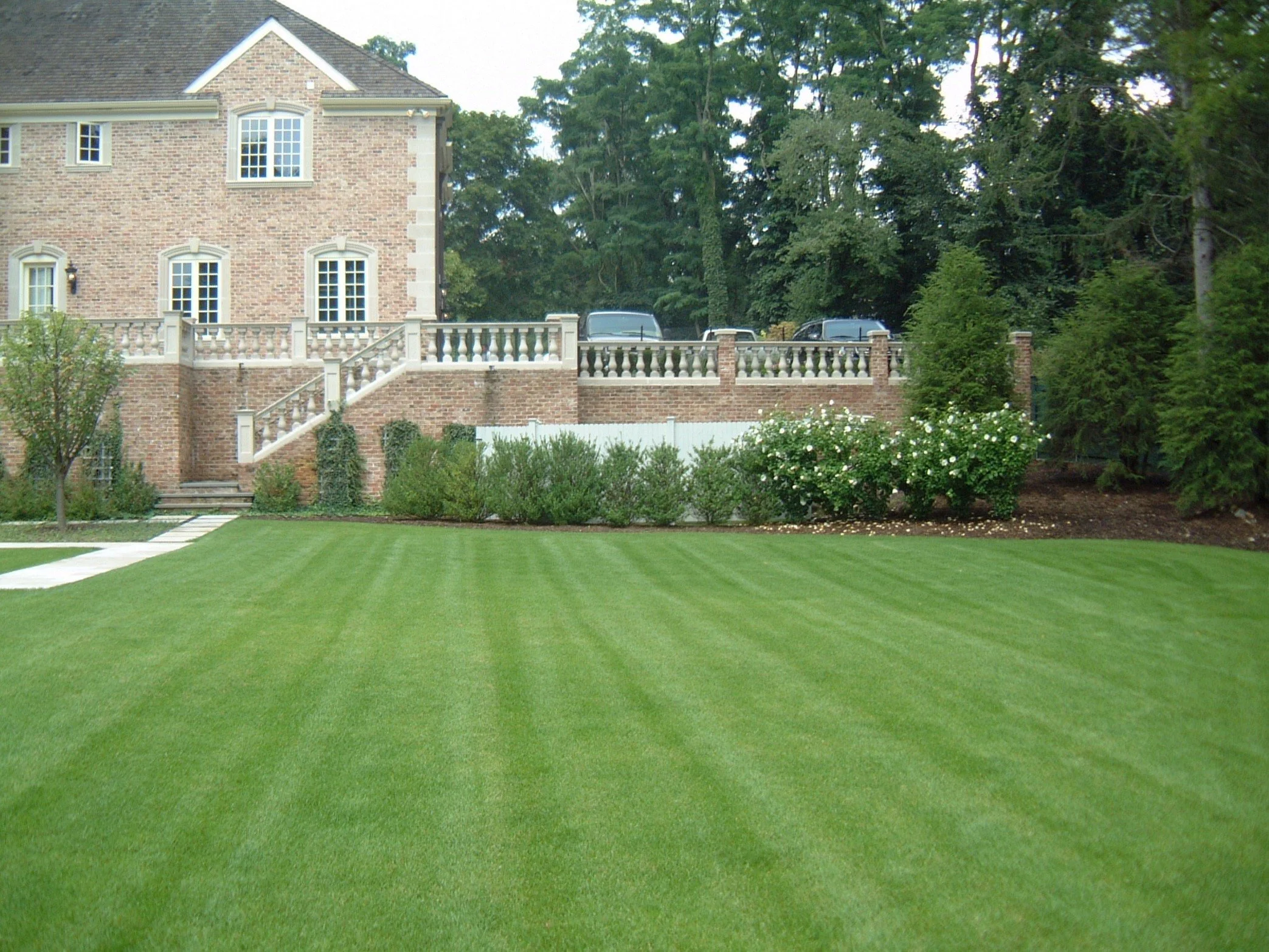 A well-maintained grassy lawn with a house and decorative brick wall in the background, surrounded by trees and landscaping.